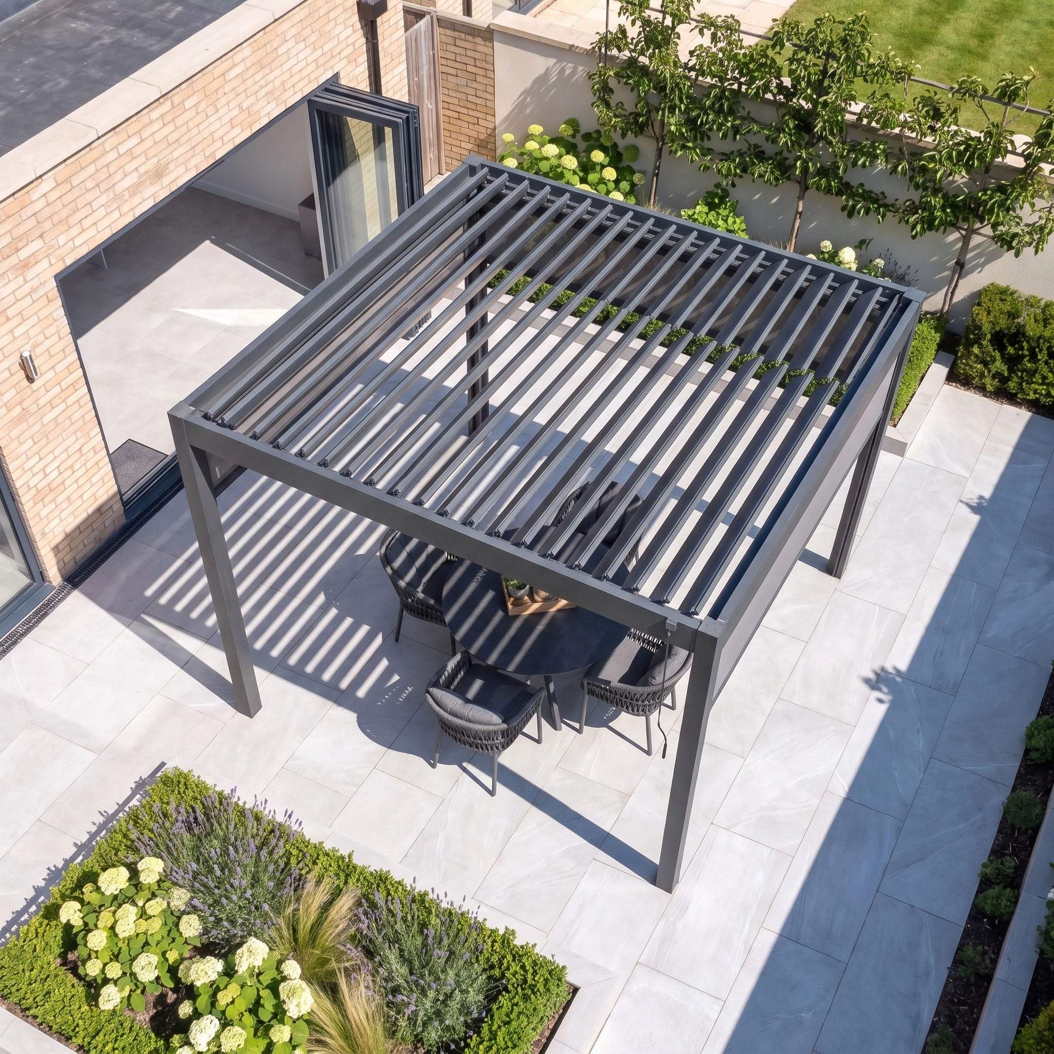 The PergoSTET Pro 3m x 3m Square Pergola with 3 Manual Side Blinds and LED Lighting in Grey features a stylish slatted roof above a patio dining set, surrounded by greenery and set on a tiled floor, providing elegant garden shade for relaxed outdoor living.