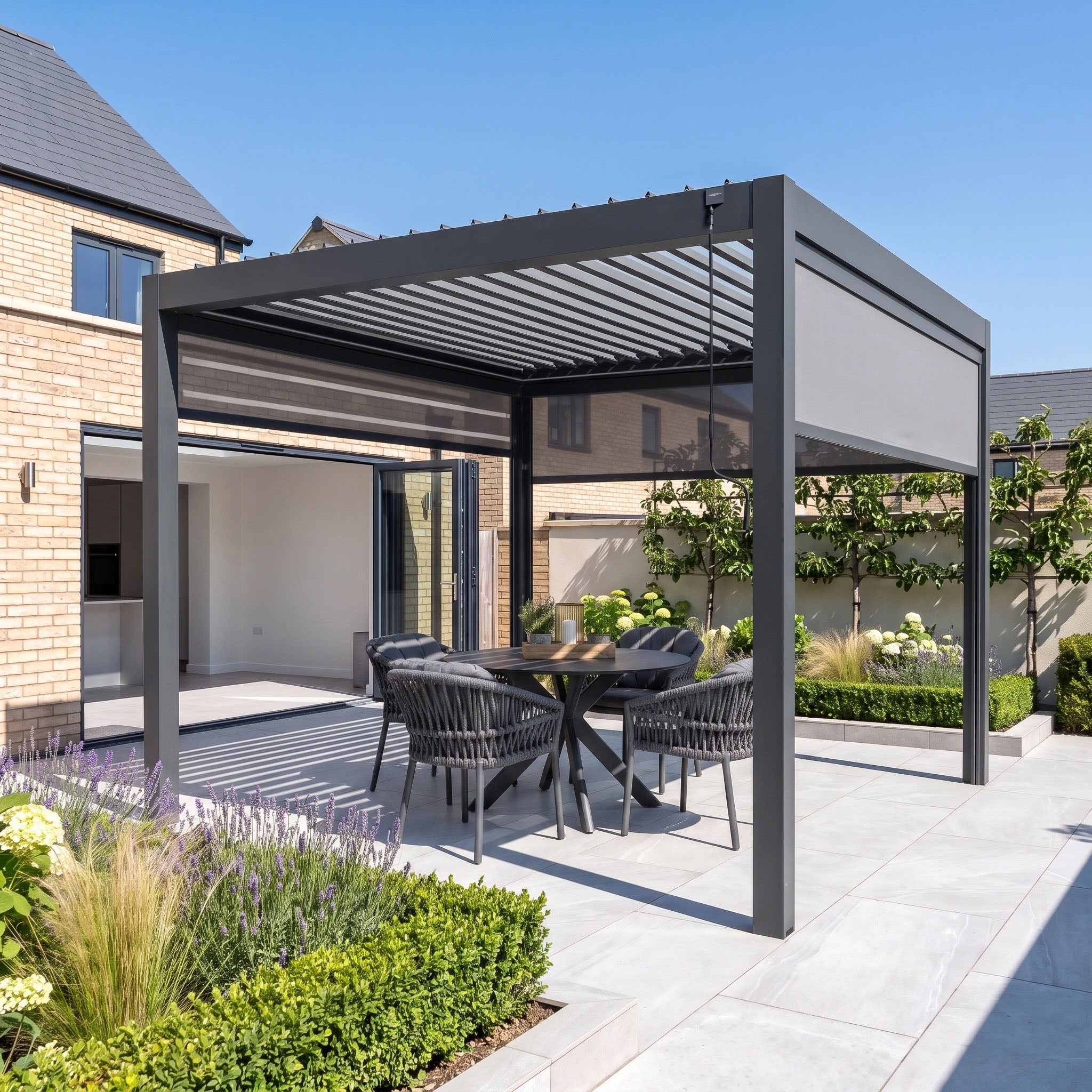 A modern patio features the PergoSTET Pro 3m x 3m Square Pergola with 3 manual side blinds and LED lighting in grey, complemented by a stylish dining table and chairs. Surrounded by lush plants and a charming brick house, this elegant aluminium pergola provides versatile garden shade and ambient illumination, making the outdoor space perfect for relaxing or entertaining.