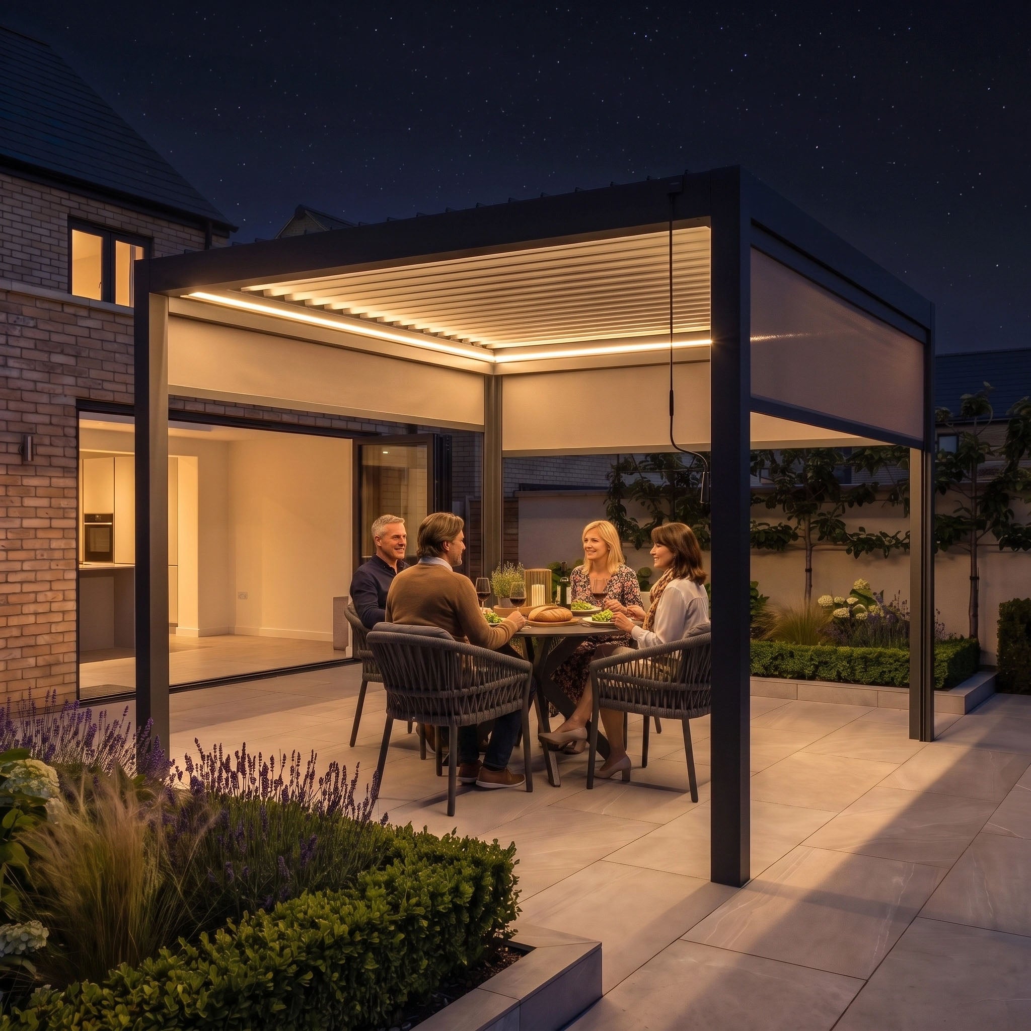 Four people dine together under the PergoSTET Pro 3m x 3m Square Pergola with 3 Manual Side Blinds and LED Lighting in Grey, enjoying a lit backyard at night surrounded by plants. This modern aluminium pergola creates an inviting atmosphere for evening gatherings.