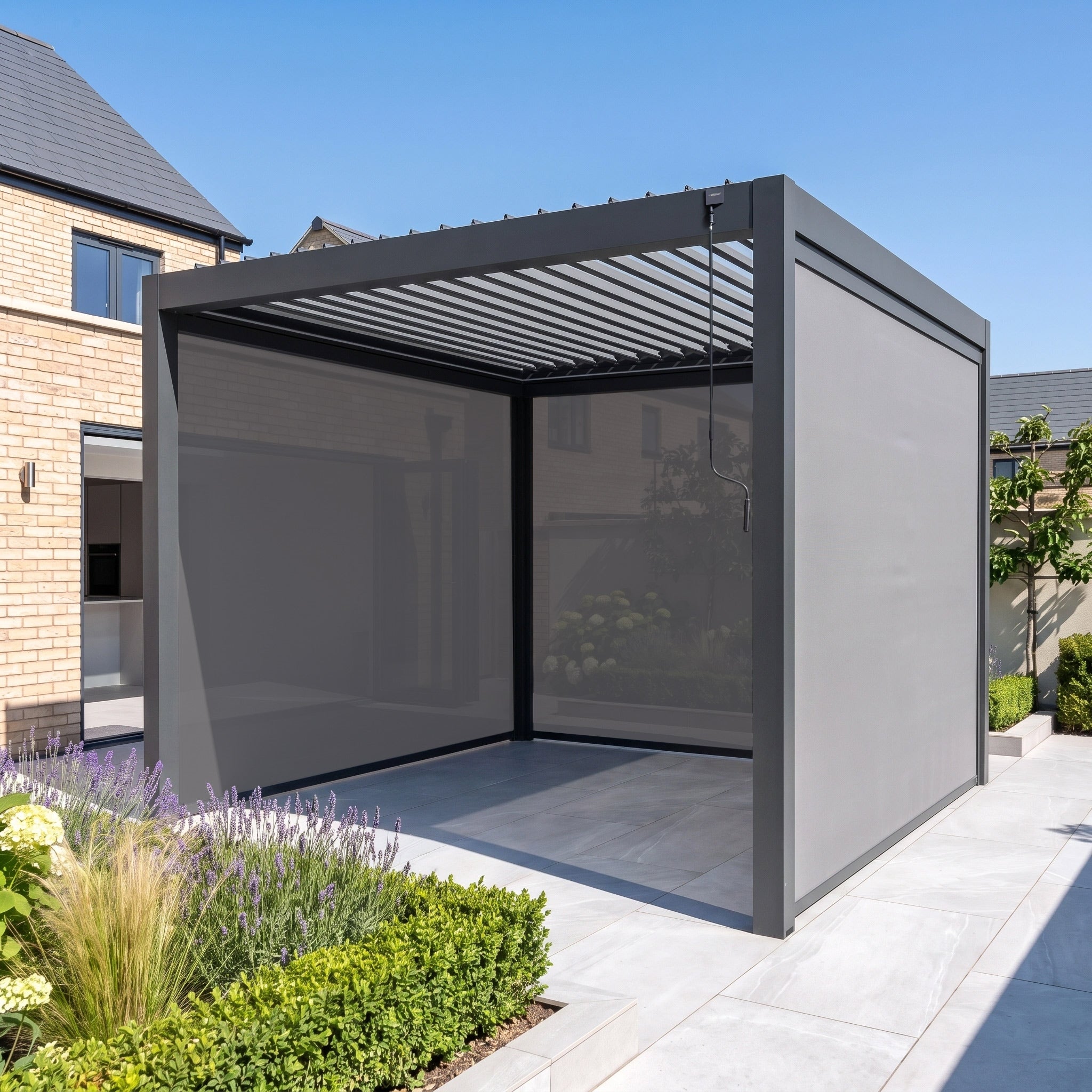 The PergoSTET Pro 3m x 3m Square Pergola, featuring a stylish grey finish, manual louvre roof, three manual side blinds, and integrated LED lighting, creates modern shade for a landscaped backyard patio.