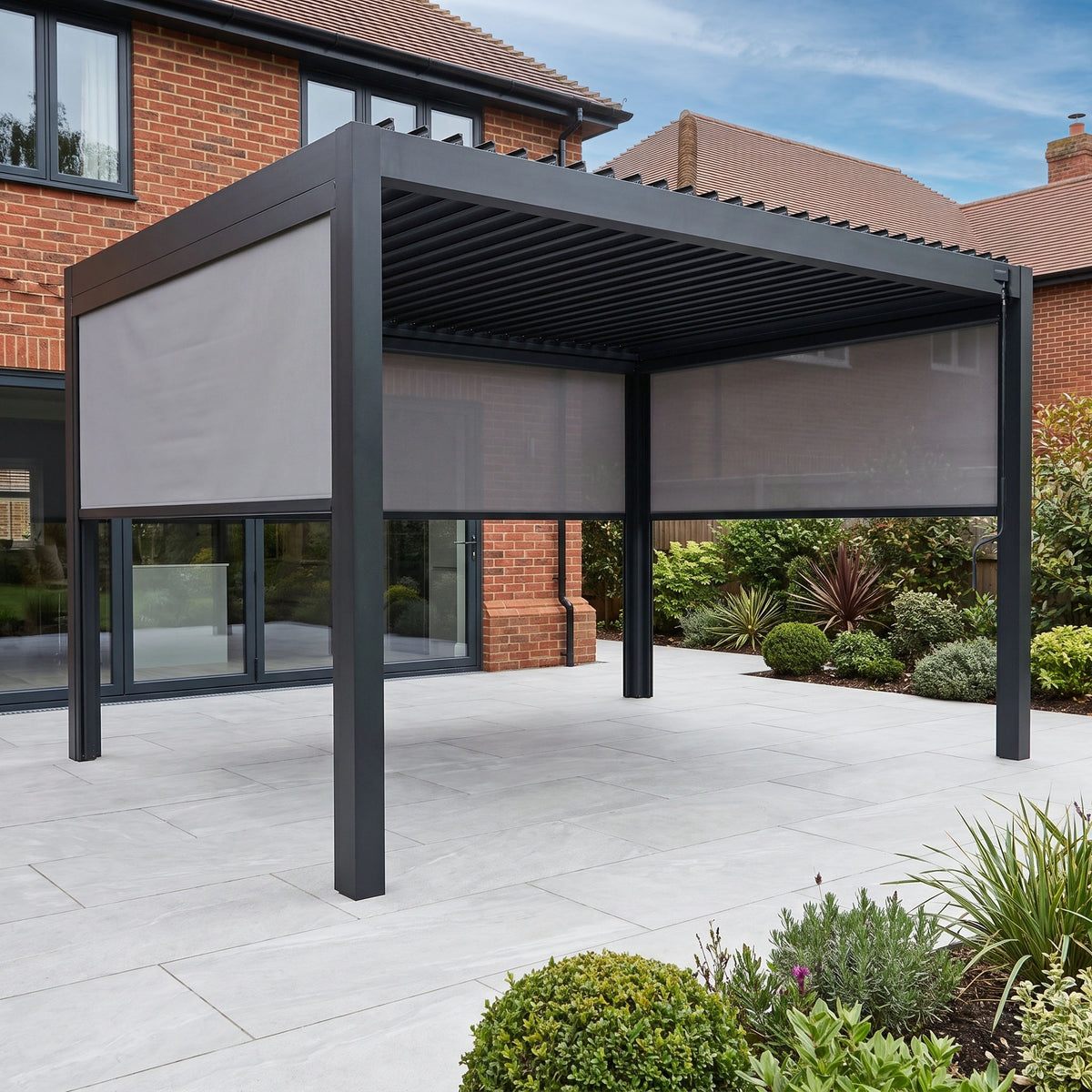 The PergoSTET Pro 3m x 4m Rectangular Pergola with 3 Manual Side Blinds and LED Lighting in Grey is installed on a tiled patio outside a brick house, surrounded by plants.