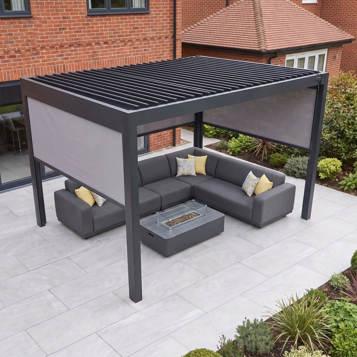 The PergoSTET Pro 3m x 4m Rectangular Pergola with 3 Manual Side Blinds and LED Lighting in Grey covers a corner sofa and fire pit table on the patio, creating a stylish and modern garden retreat that’s perfect for relaxing outdoors.
