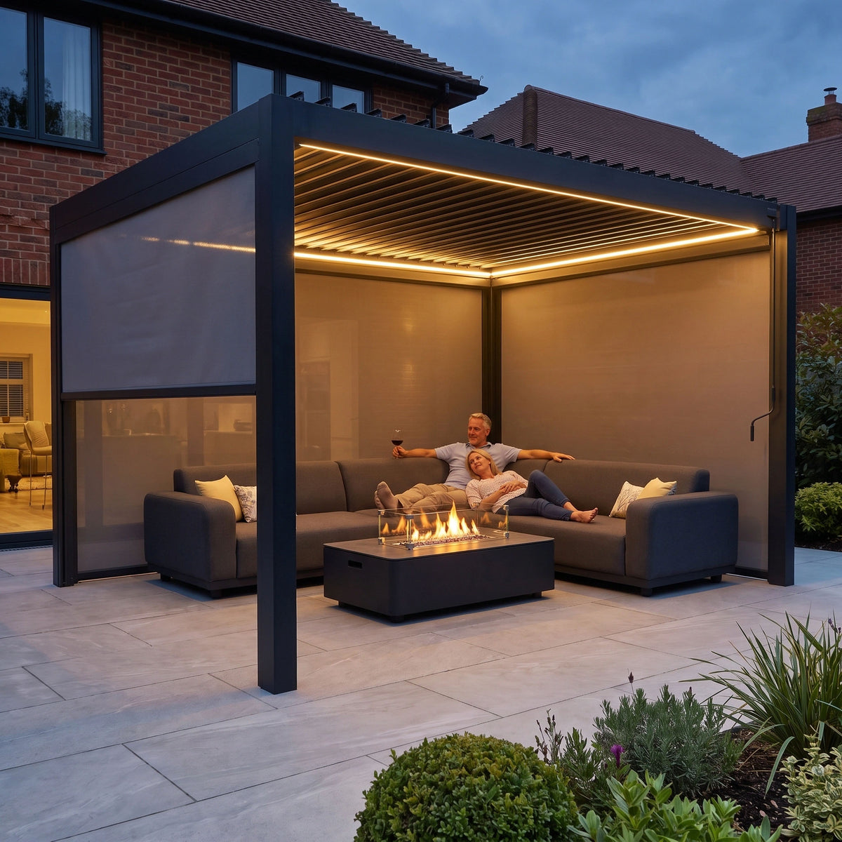 A couple relaxes by a fire table under the PergoSTET Pro 3m x 4m Rectangular Pergola with 3 Manual Side Blinds and LED Lighting in Grey in their backyard at dusk.