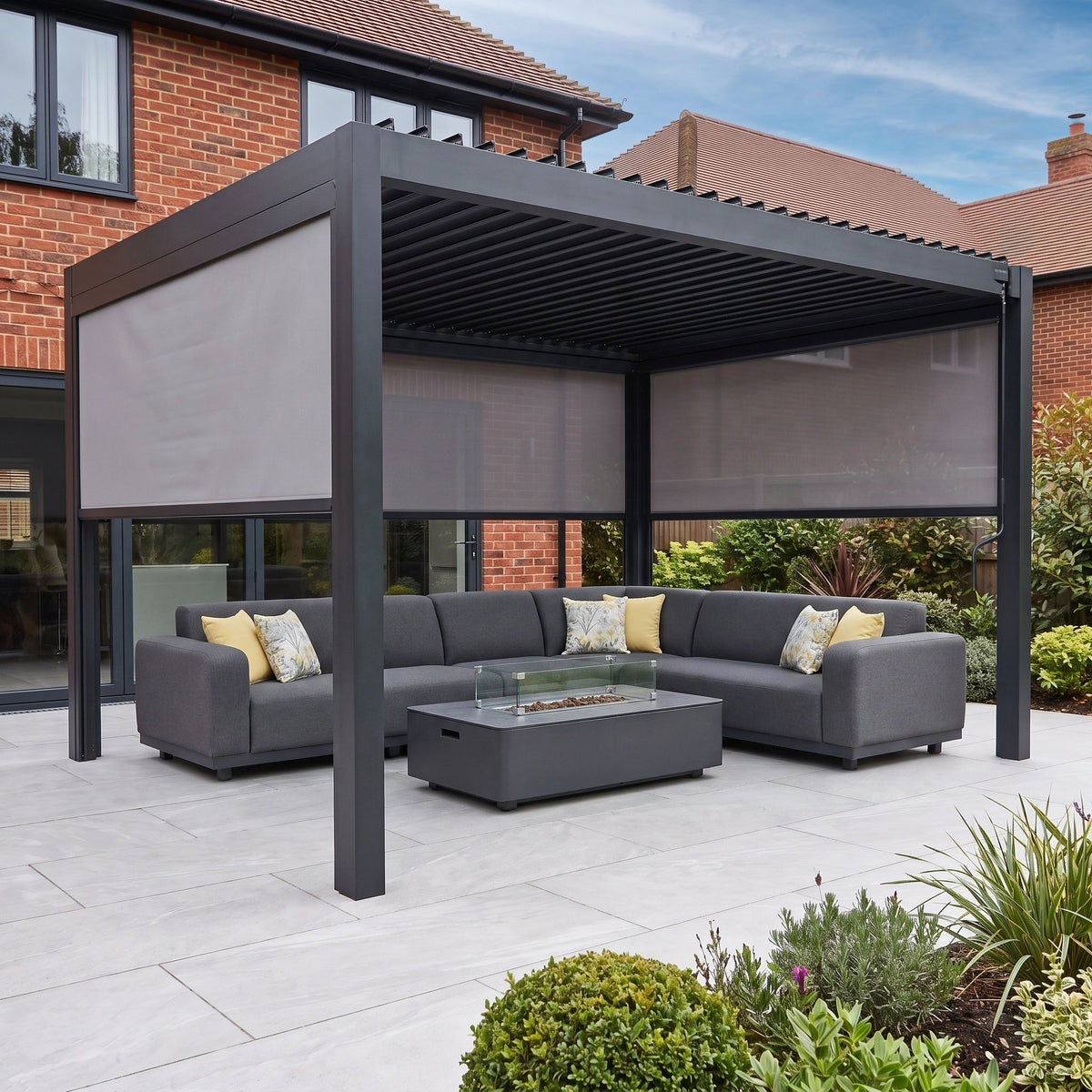 The PergoSTET Pro 3m x 4m Rectangular Pergola with 3 Manual Side Blinds and LED Lighting in Grey creates an inviting outdoor retreat when paired with gray sectional sofas, a fire table, and lush surrounding plants on the patio. Its sleek aluminium design blends seamlessly with a contemporary setting, offering both style and adjustable shade.