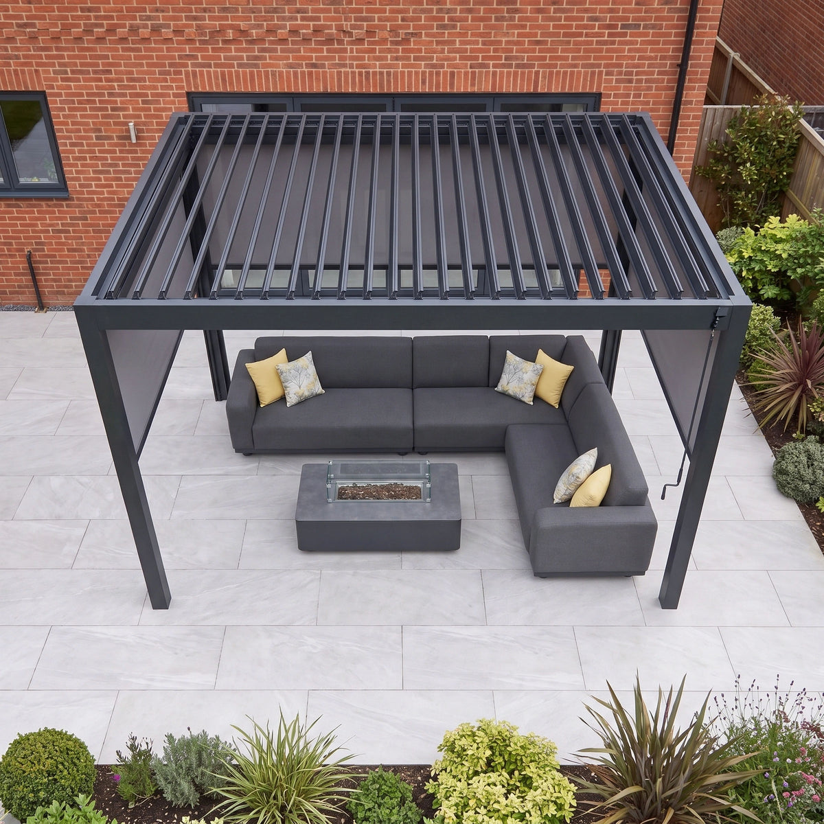 A modern outdoor patio featuring a gray sectional sofa, yellow pillows, and the PergoSTET Pro 3m x 4m Rectangular Pergola with 3 manual side blinds and LED lighting in grey on a tiled floor.