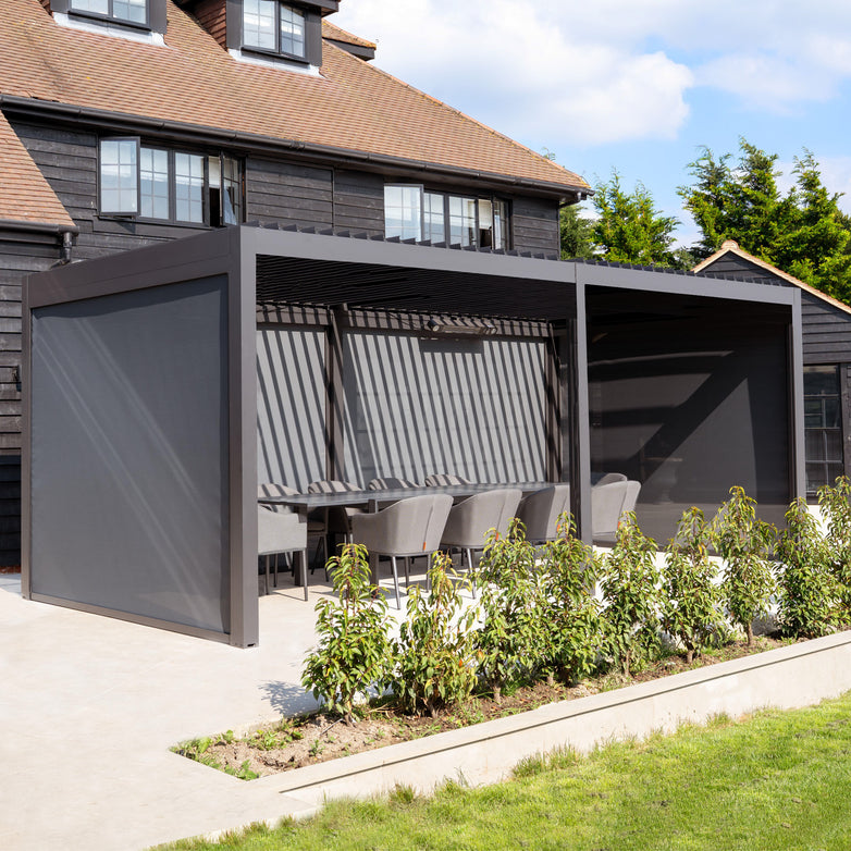 The PergoSTET Deluxe 3m x 6m Rectangular Pergola in grey features motorised LED louvres and 4 manual side blinds, set with dining chairs and table beside a black house, surrounded by greenery.