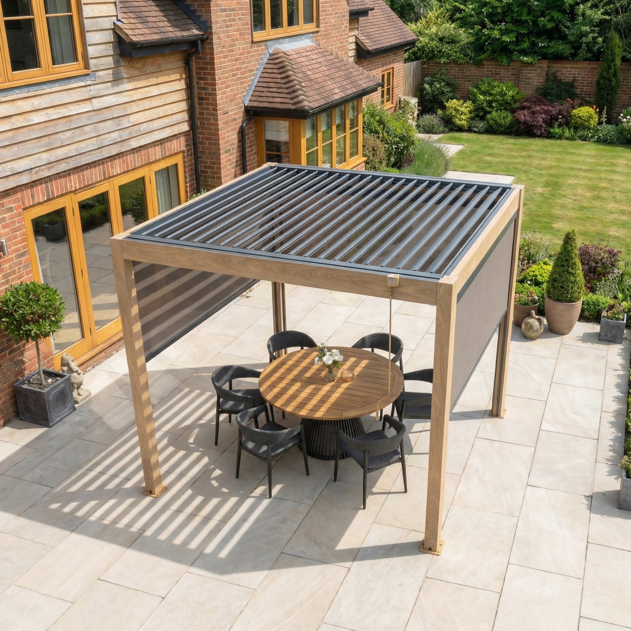 The PergoSTET Plus 3m x 3m Square Pergola with 3 manual side blinds and LED lighting in wood effect shelters a round table and chairs on a landscaped patio.
