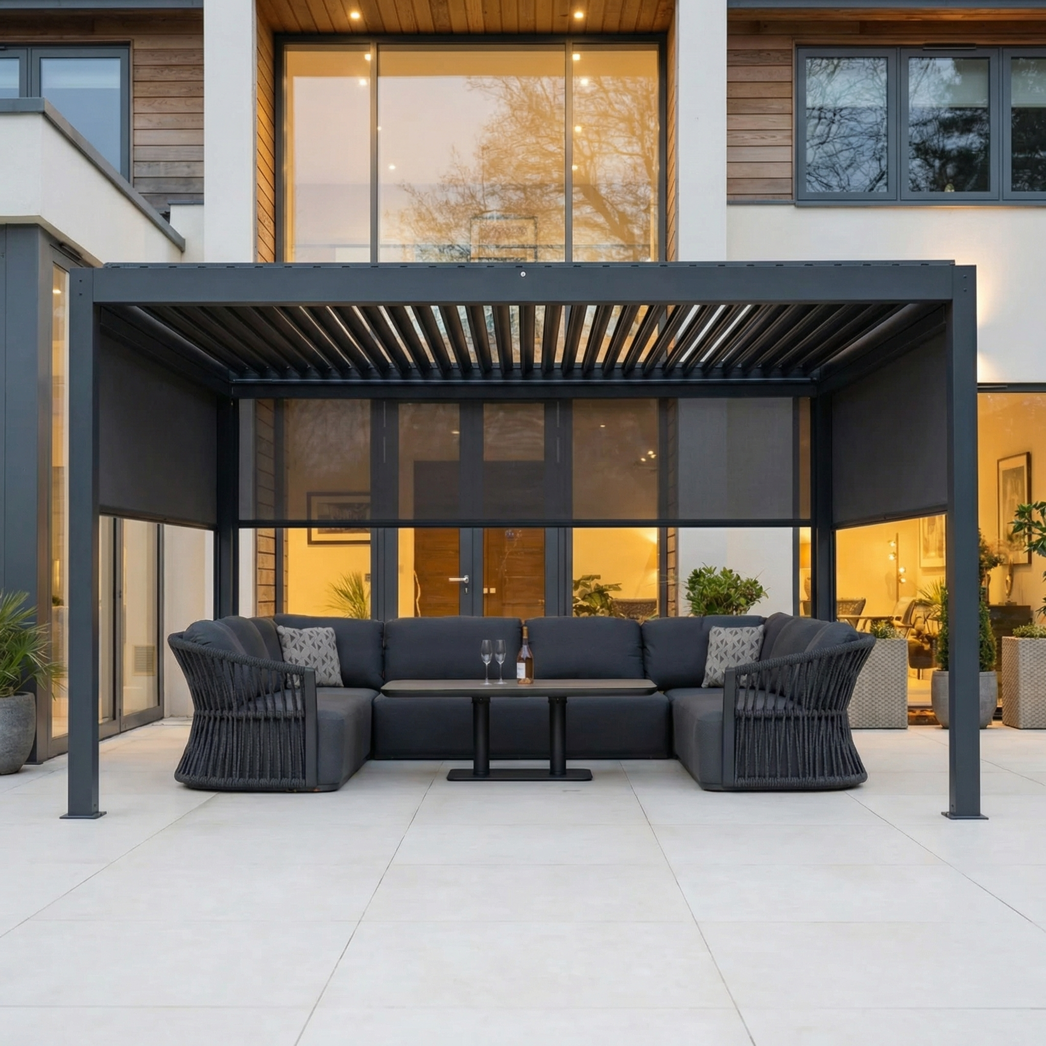 Contemporary outdoor seating set with sofa and armchairs arranged beneath a PergoSTET Plus 3m x 4m Rectangular Pergola featuring 3 manual side blinds and LED lighting in grey, placed in front of a house.