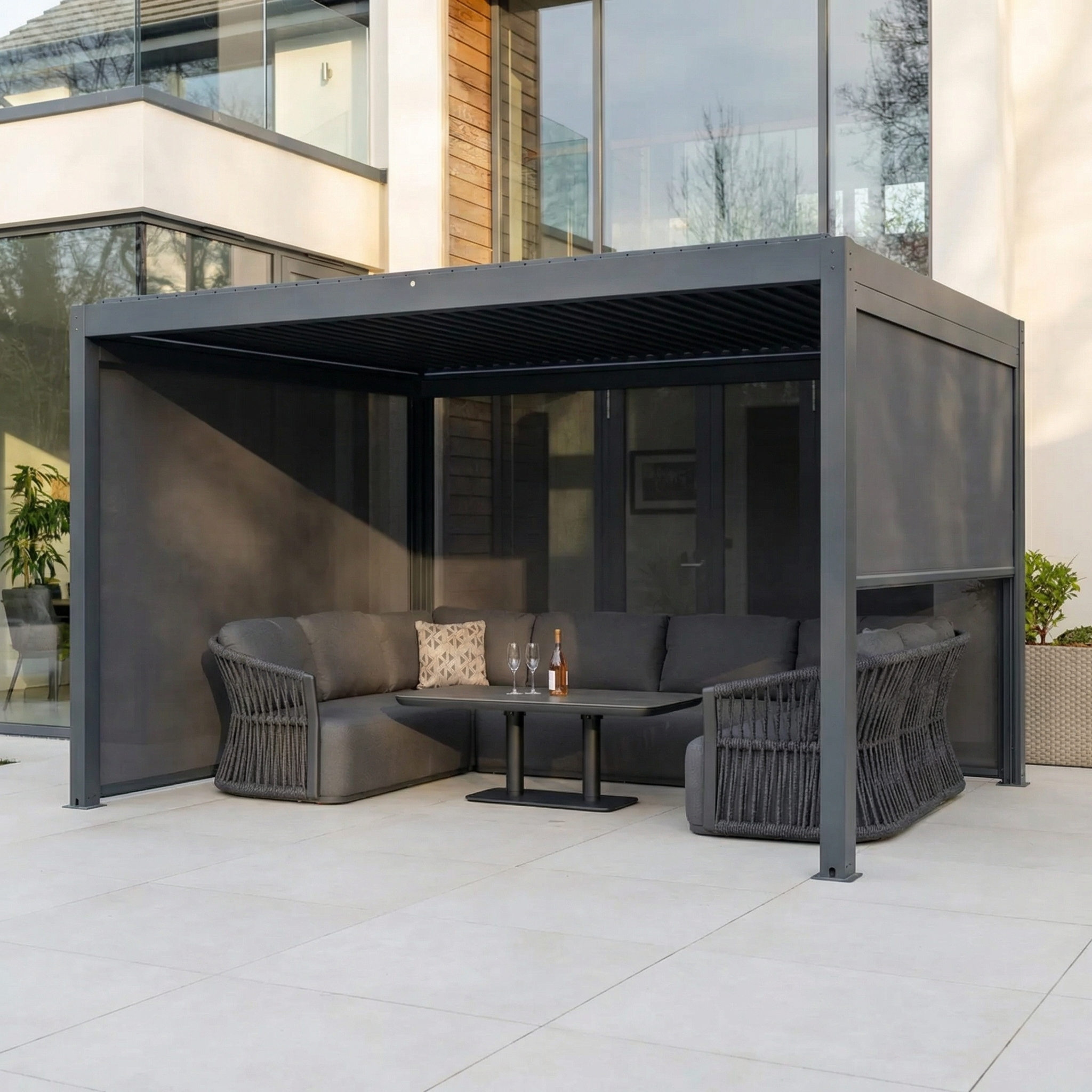 The spacious patio features the PergoSTET Plus 3m x 4m Rectangular Pergola with 3 manual side blinds and LED lighting in grey, complemented by a gray sectional sofa, table, and drinks by the house.