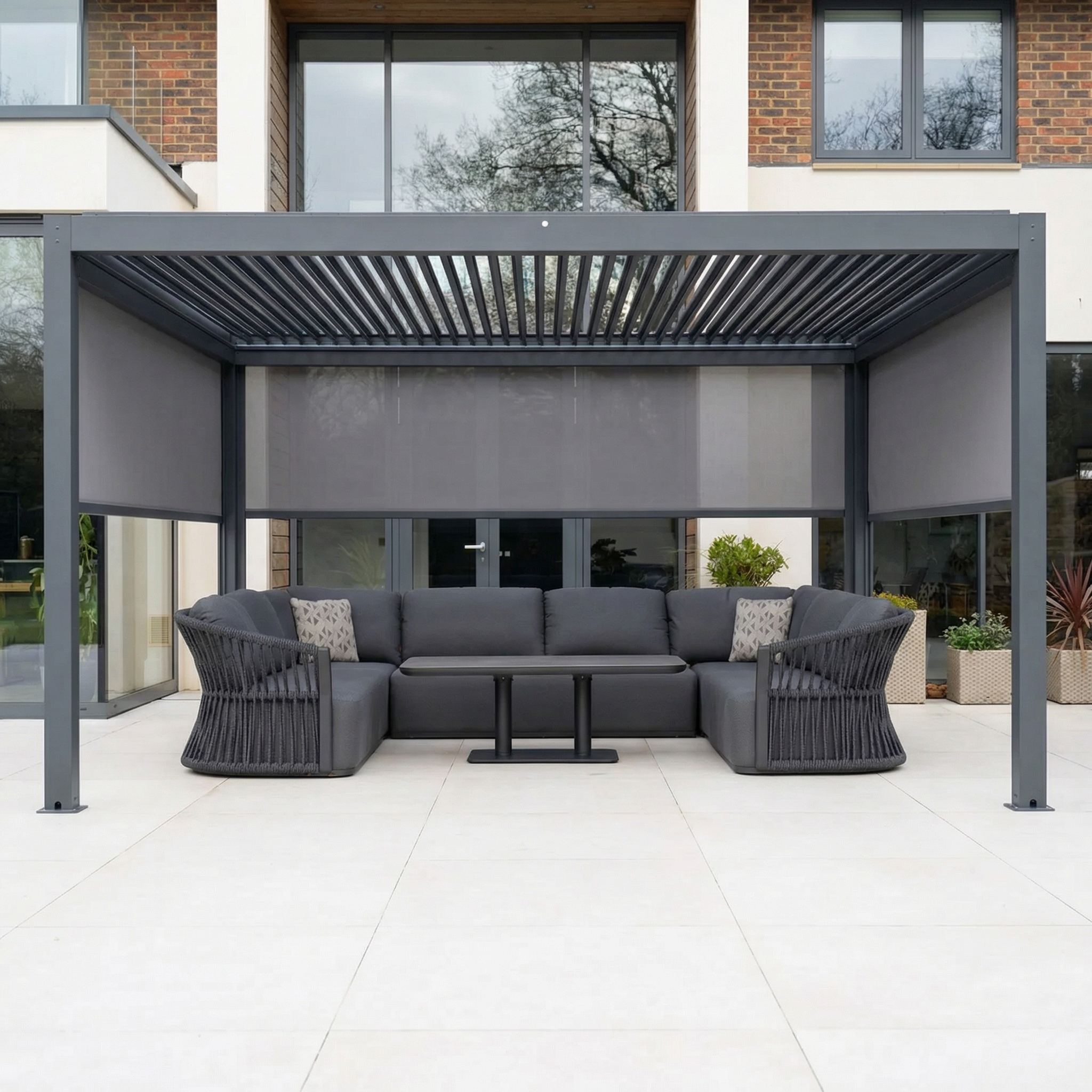 The PergoSTET Plus 3m x 4m Rectangular Pergola in Grey features 3 manual side blinds and LED lighting, shown with a gray sectional sofa, armchairs, and table on a tiled patio in front of the house.