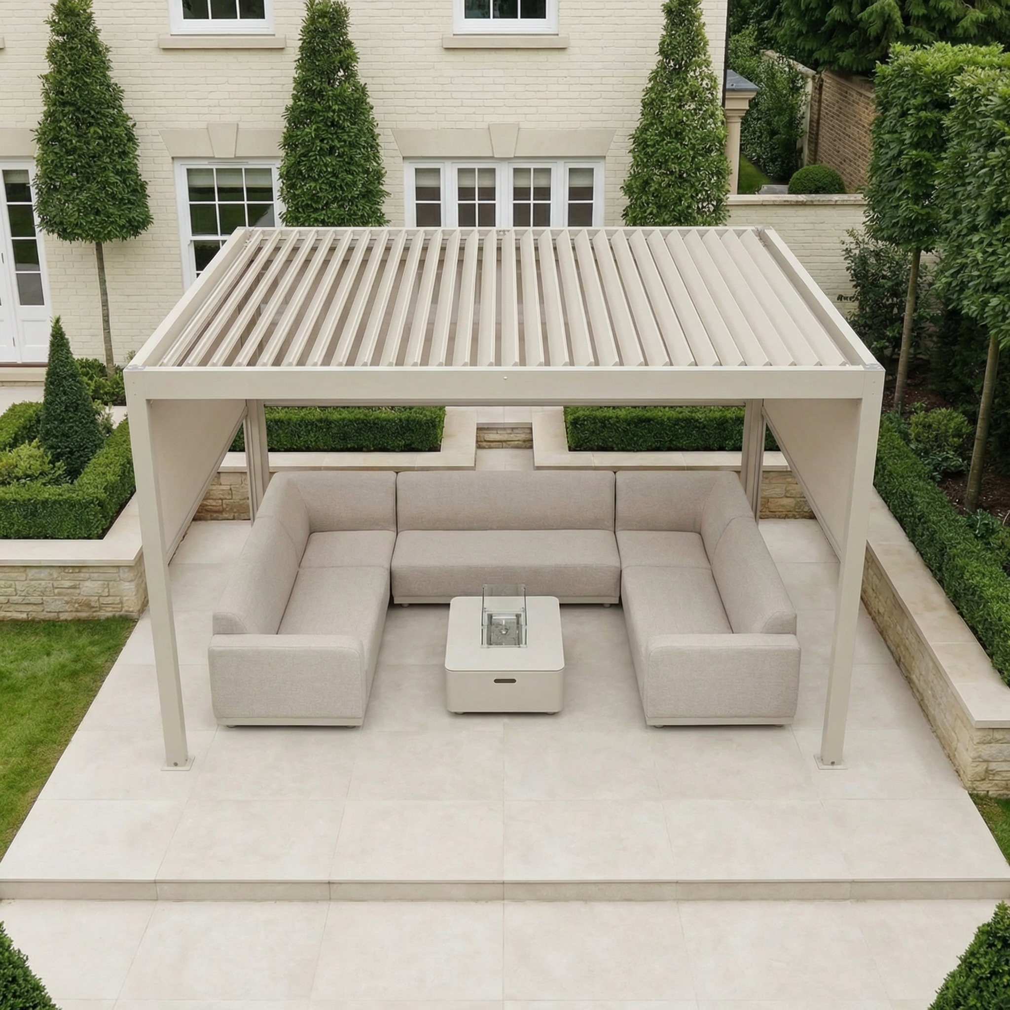 Modern outdoor patio with cream sofas and table under a PergoSTET Plus 3m x 4m Rectangular Pergola in Latte, featuring 3 manual side blinds and LED lighting, surrounded by neatly trimmed greenery for a stylish setting.