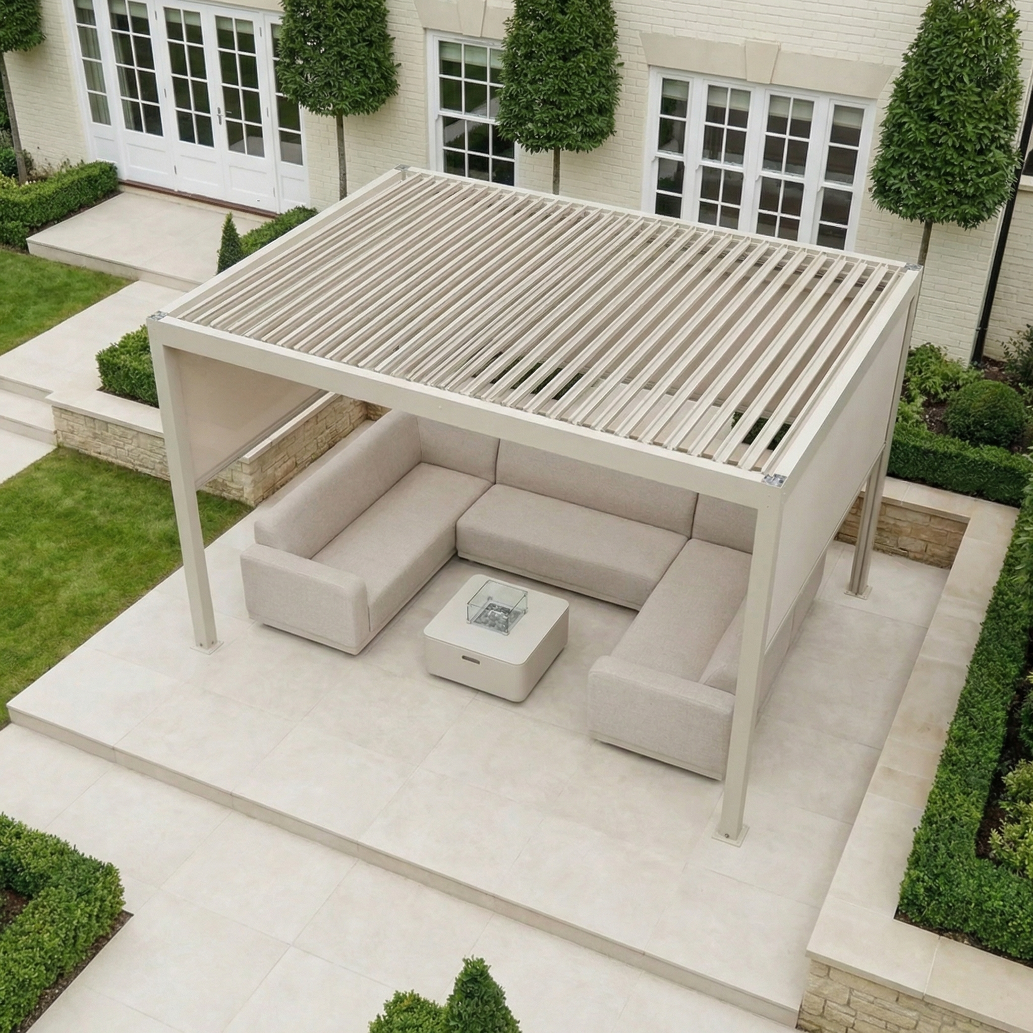The PergoSTET Plus 3m x 4m Rectangular Pergola with three manual side blinds and LED lighting in Latte creates a cozy patio, complemented by beige sectional sofas, a fire pit table, and lush greenery for an inviting outdoor retreat.