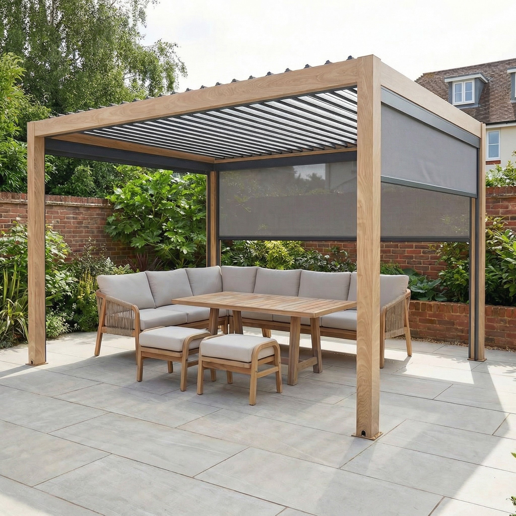 The PergoSTET Plus 3m x 4m wood-effect pergola with 3 manual side blinds and LED lighting adds stylish comfort to a paved backyard patio, paired with beige cushioned seating, a table, and lush greenery.