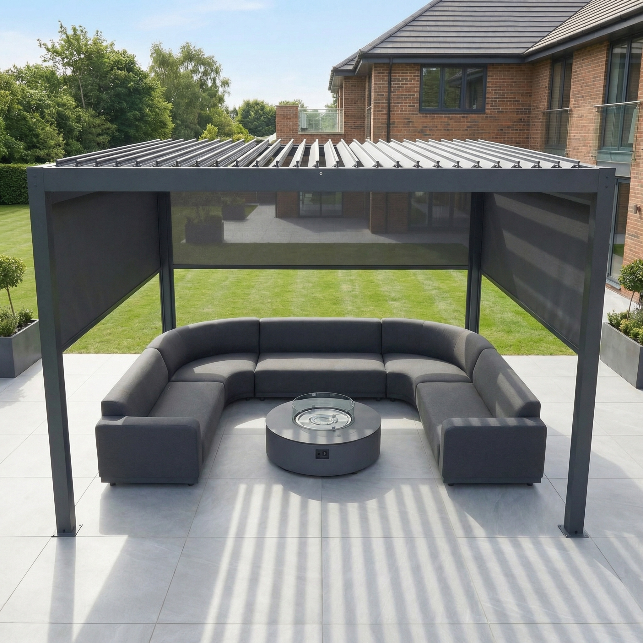 The PergoSTET Plus 4m x 4m Square Pergola with 3 Manual Side Blinds and LED Lighting in Grey frames a curved gray sofa and fire pit on a tiled patio beside the lawn and house.
