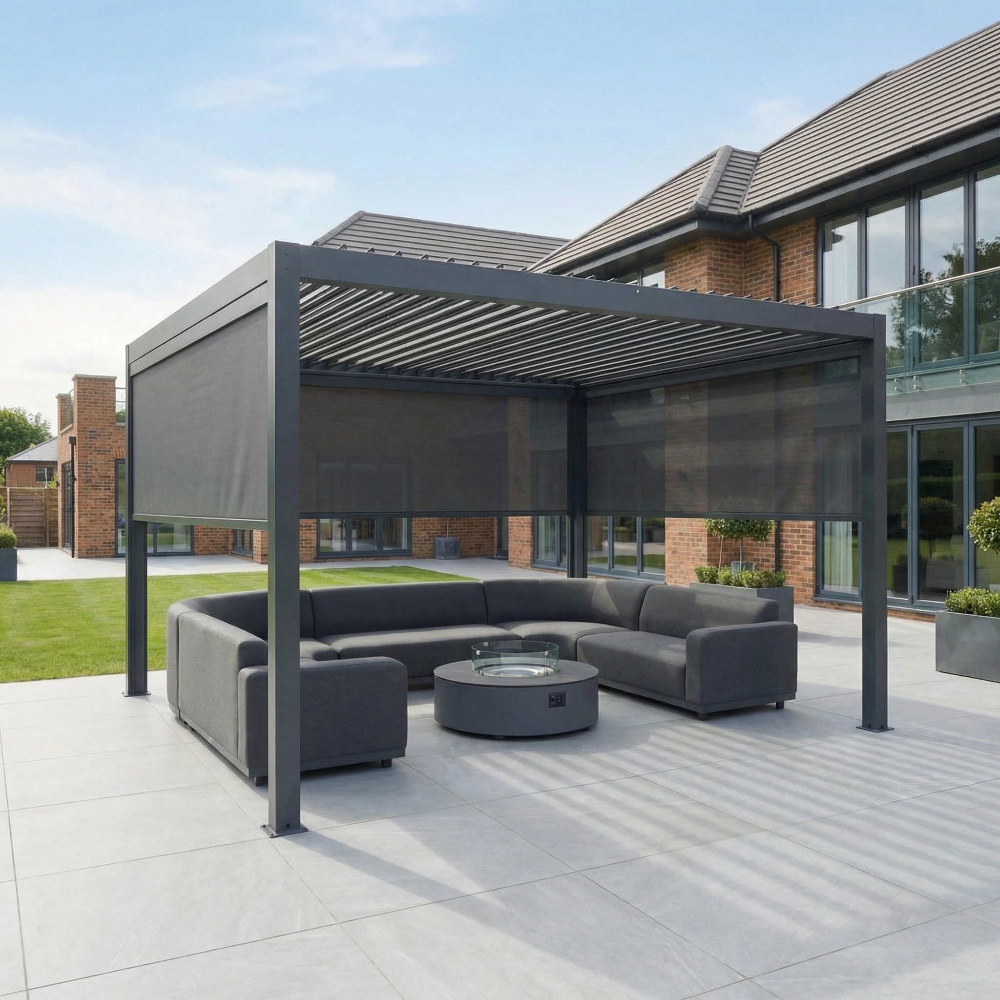 Modern patio featuring the PergoSTET Plus 4m x 4m square pergola in grey with LED lighting and 3 manual side blinds, a sectional sofa, fire pit table, and a large brick house in the background.