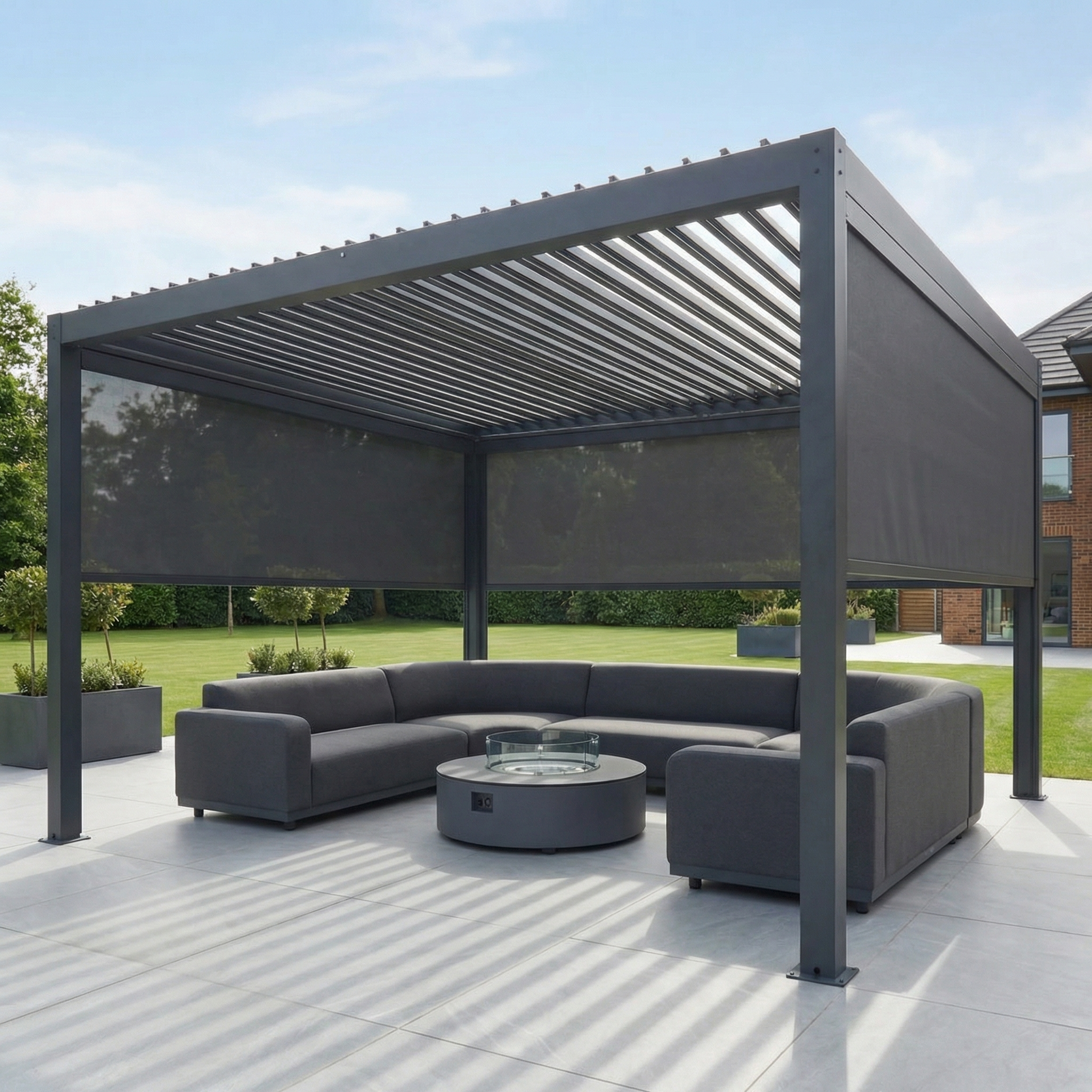 PergoSTET Plus 4m x 4m Square Pergola with 3 manual side blinds and LED lighting in grey, featuring a gray sectional sofa and round fire pit on a patio, surrounded by grass and trees.