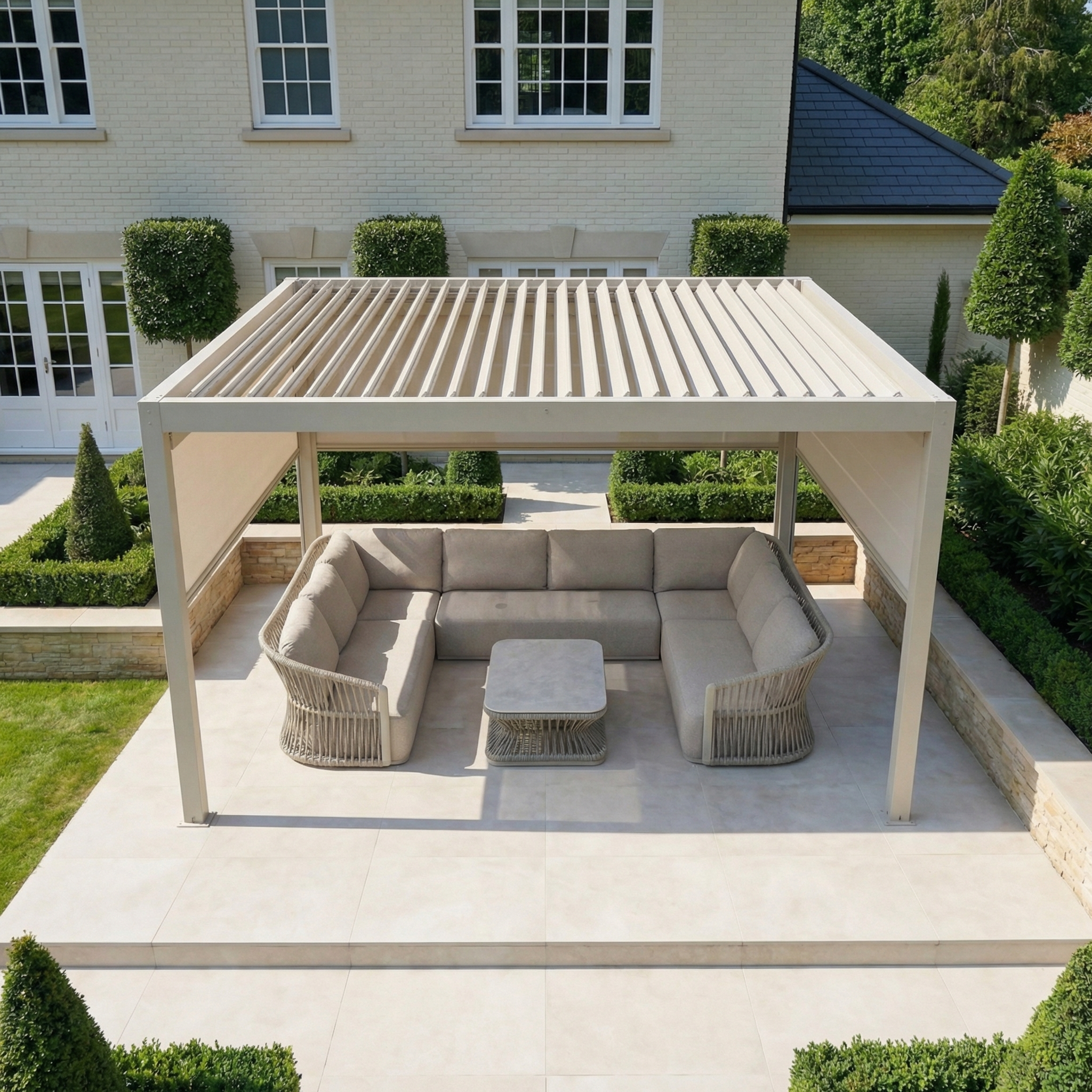 Modern patio with the PergoSTET Plus 4m x 4m Square Pergola in Latte, featuring tilting louvres, LED lighting, and 3 manual side blinds, complemented by beige sofas and a square table in a landscaped garden.