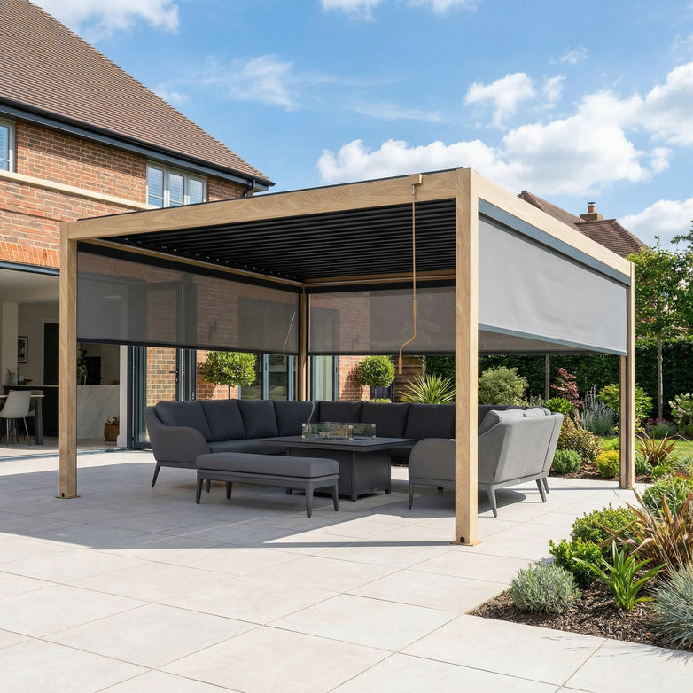 The PergoSTET Plus 4m x 4m Square Pergola with manual blinds and LED lighting in wood effect frames a gray sectional sofa and table on the tiled patio beside the brick house.