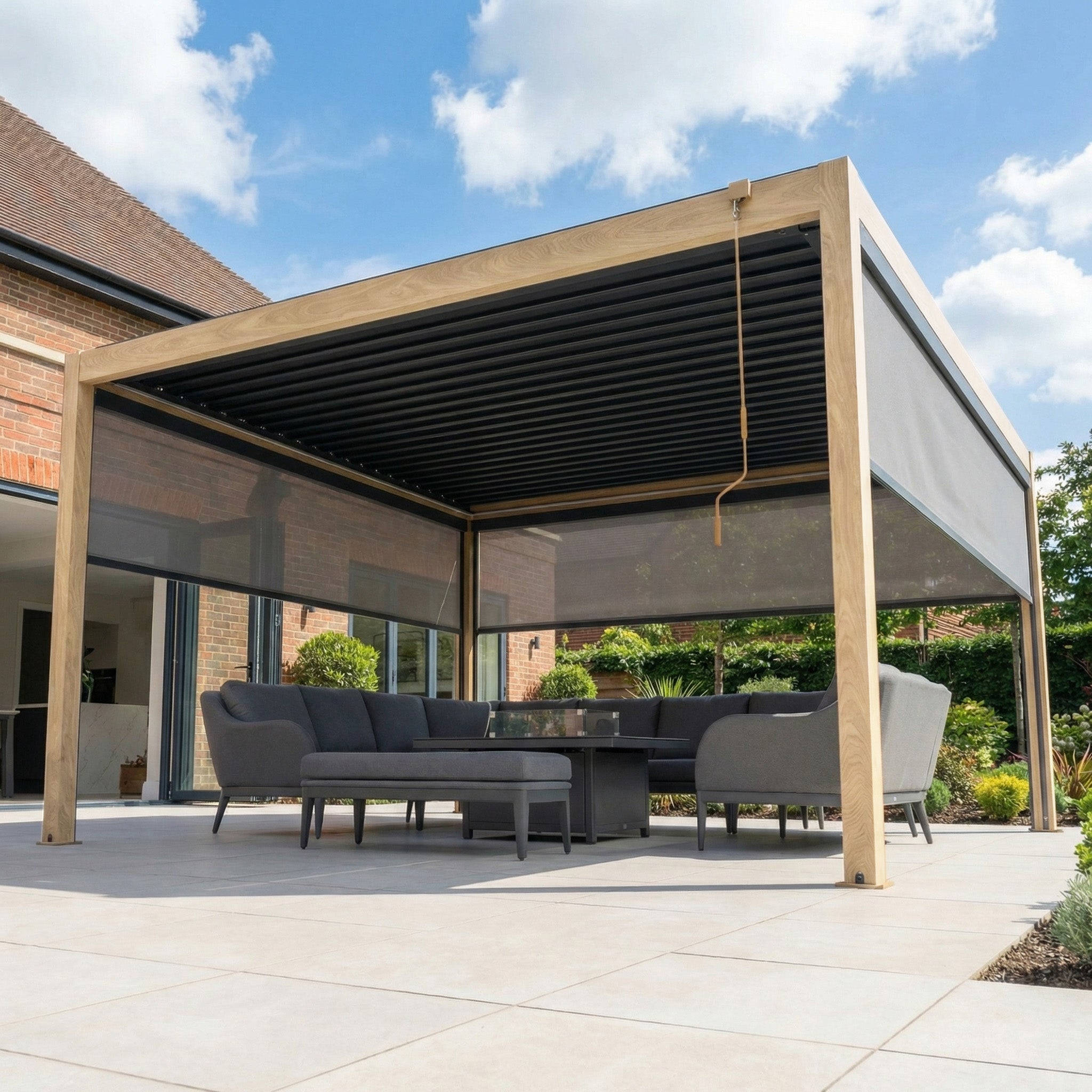 The PergoSTET Plus 4m x 4m Square Pergola in wood effect features a modern frame, LED lighting, and comes with 3 manual side blinds for added style and comfort—perfect for patios beside brick houses.
