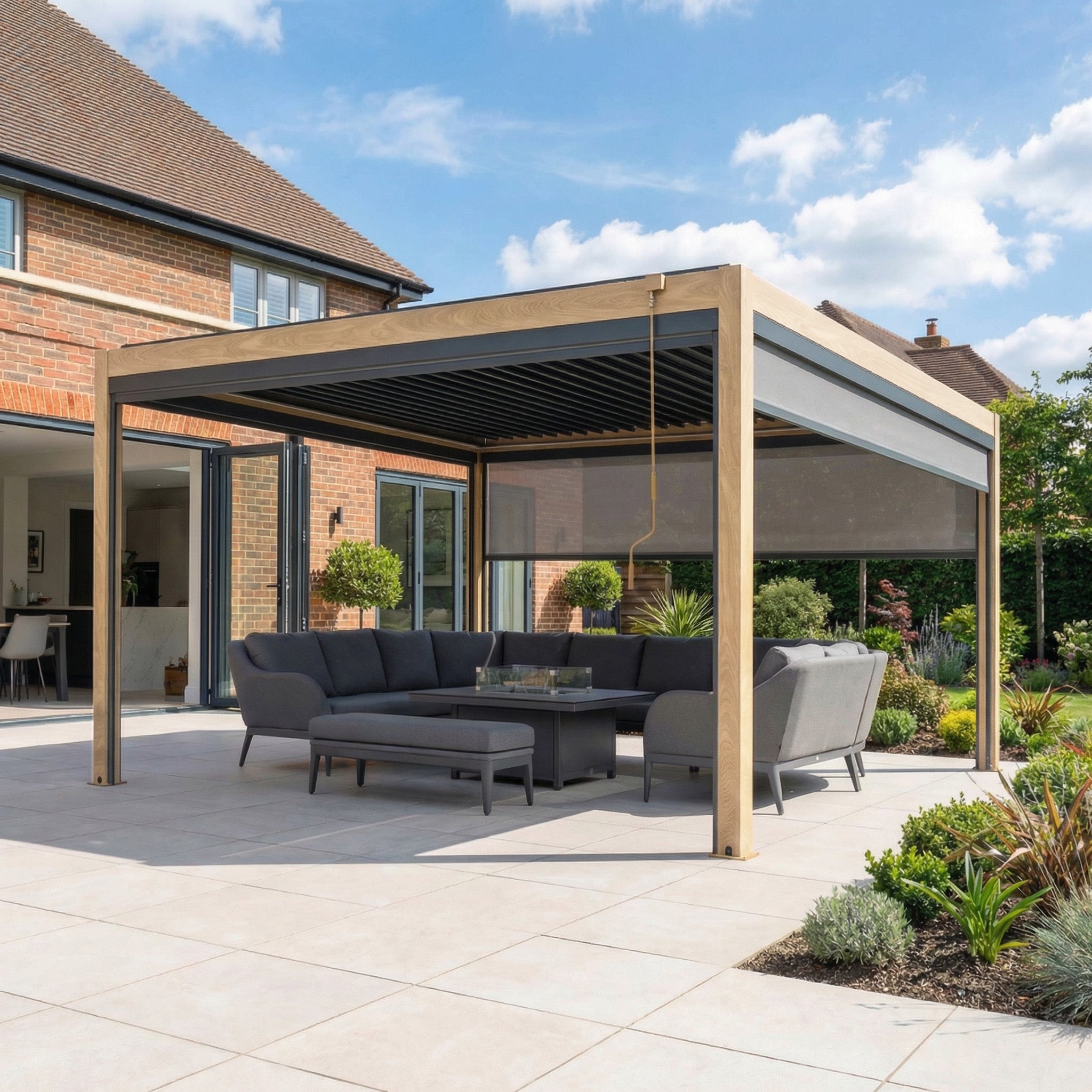 The PergoSTET Plus 4m x 4m Square Pergola with 3 manual side blinds and LED lighting in wood effect shades a gray outdoor sofa set on a tiled patio beside a brick house and garden.