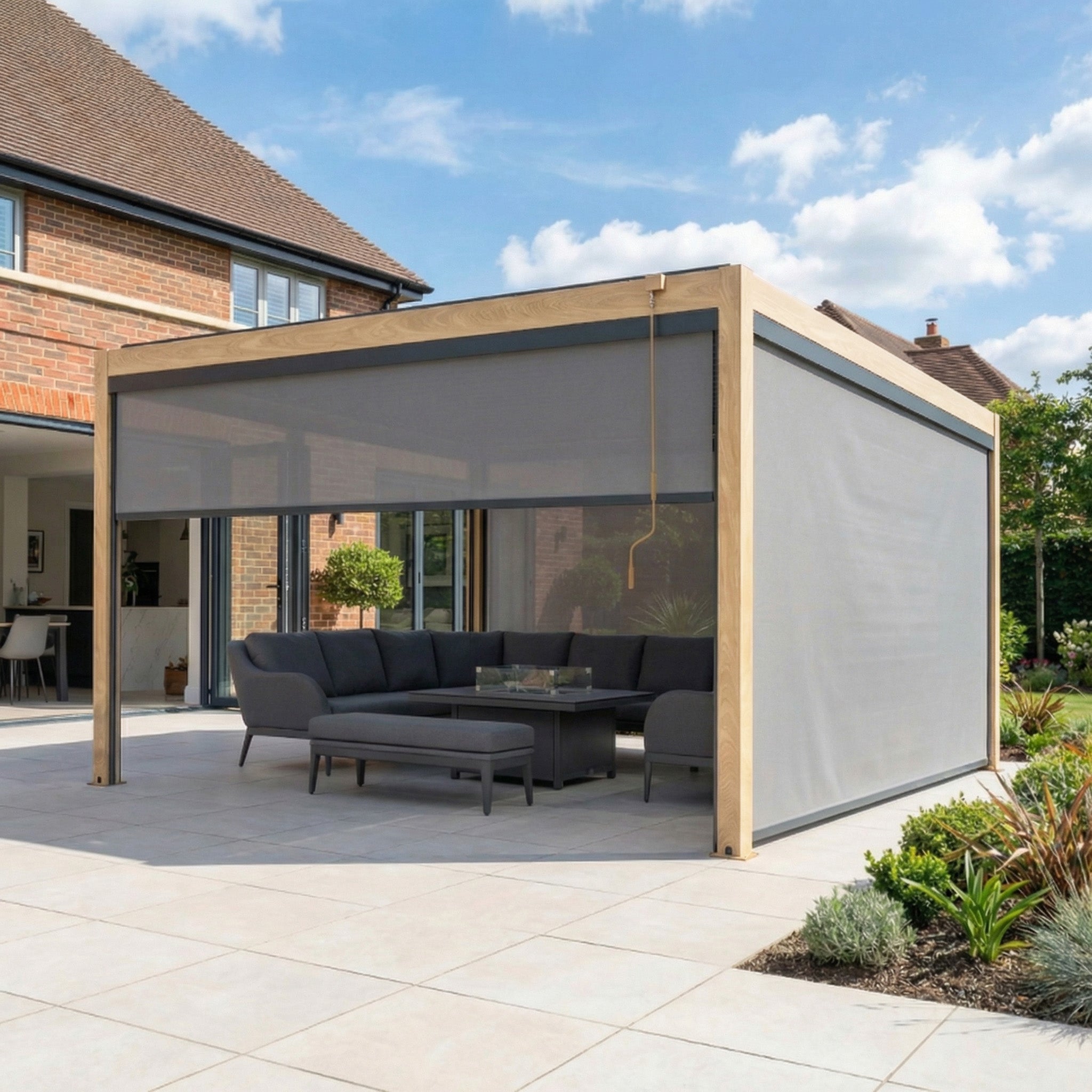 The PergoSTET Plus 4m x 4m Square Pergola with 3 manual side blinds and LED lighting in wood effect frames a gray seating area and coffee table on the patio, creating a stylish outdoor retreat.