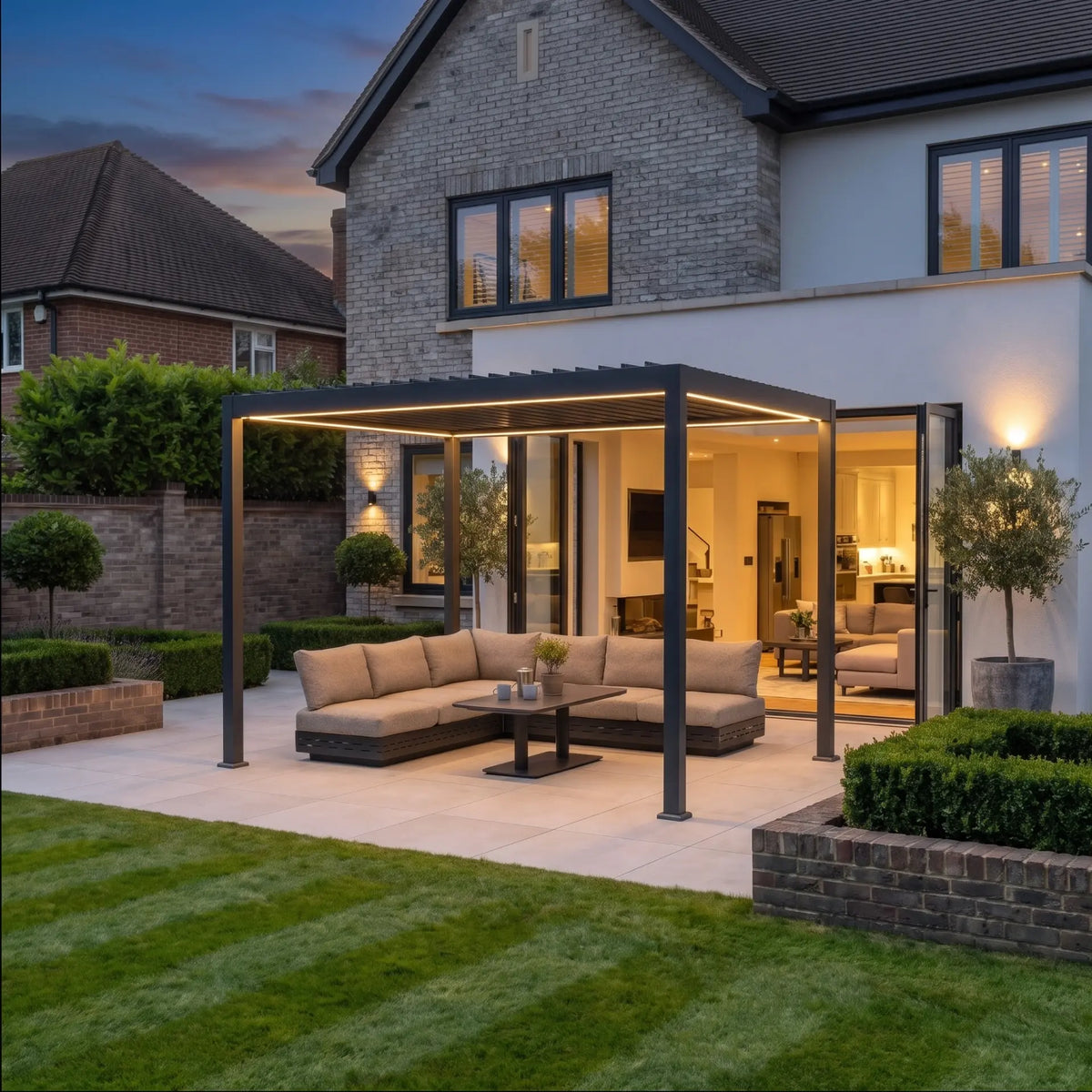 A modern patio features the PergoSTET Lite 3m x 4m Metal Rectangular Pergola With Motorised Louvres and LED Lighting in Grey, sheltering an outdoor sofa set beneath its solar-powered structure. Warm LED lights illuminate the space, complementing the contemporary house at dusk.