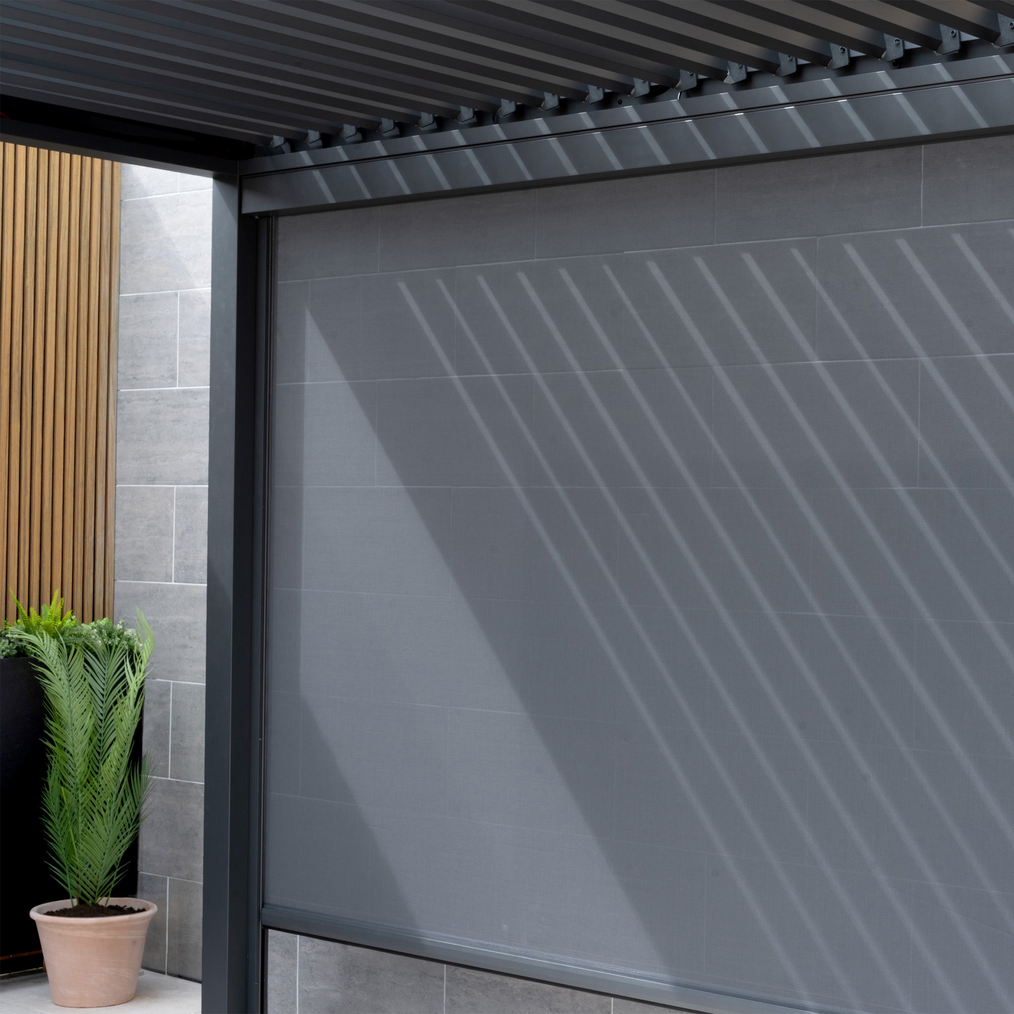 A modern patio showcases a sleek PergoSTET Deluxe 3m x 6m Rectangular Pergola with motorised LED louvres and four manual side blinds in grey, casting striped shadows on a gray wall beside a planter with lush green foliage.