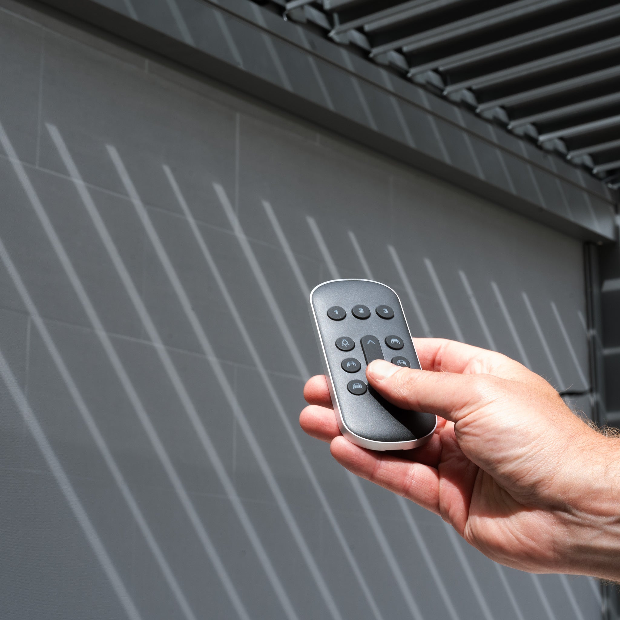 A hand grips a remote to maneuver the PergoSTET Deluxe 3m x 6m Pergola, casting intricate shadows on the wall with its motorised LED louvres.