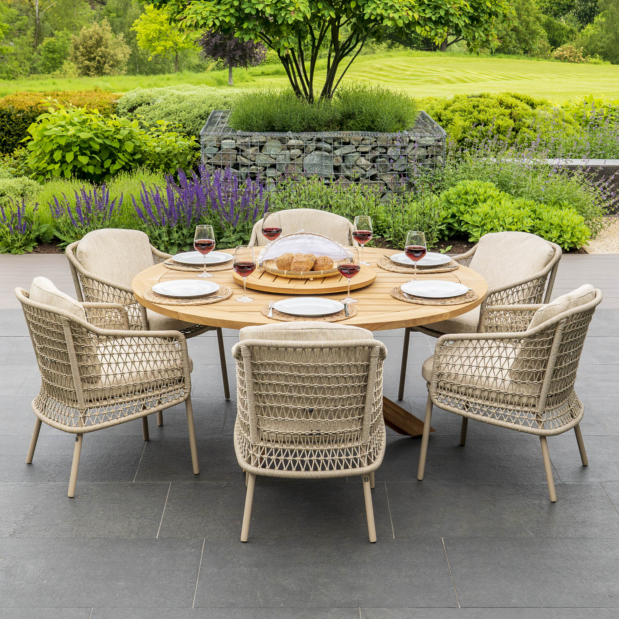 Outdoor dining setup with a Puccini 6 Seat Round Dining Set with Lazy Susan in Latte, featuring a teak wood table, six wicker chairs from the 4 Seasons Rope collection, wine glasses, plates, and a bread dish, all set in a lush garden.