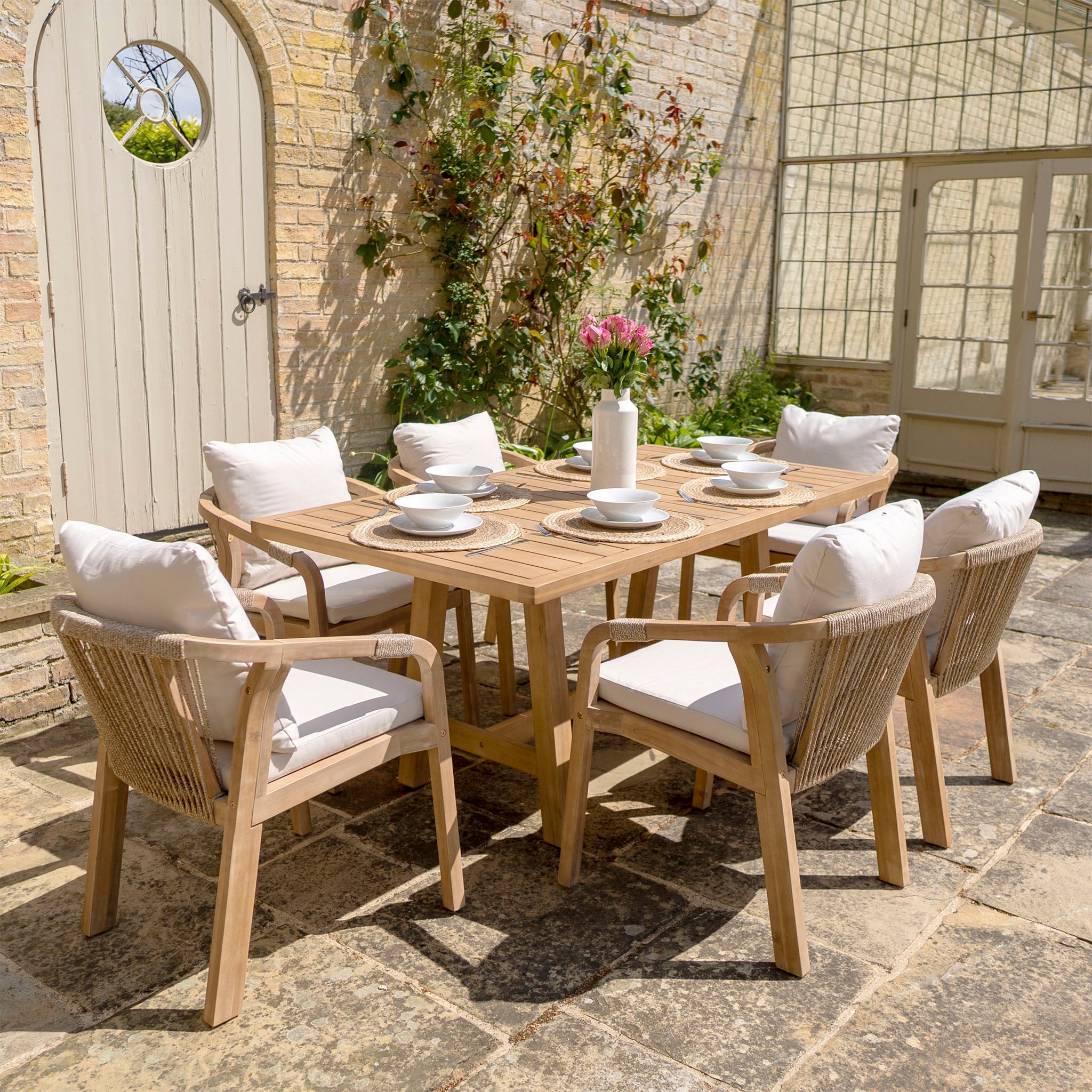 A patio featuring the Quay 6 Seat Rectangular Dining Set in Linen, crafted from sustainably sourced acacia wood, with six cushioned chairs upholstered in water-resistant fabric, and a rectangular table set for a meal, all surrounded by a lush garden.