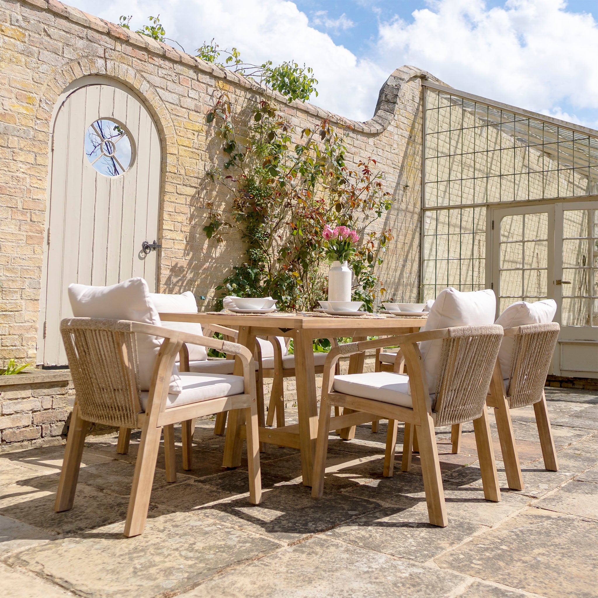 Outdoor patio featuring the Quay 6 Seat Rectangular Dining Set in Linen, crafted from sustainably sourced acacia wood, cushioned chairs made of water-resistant fabric, a stone wall adorned with climbing plants, and a round-topped wooden door.
