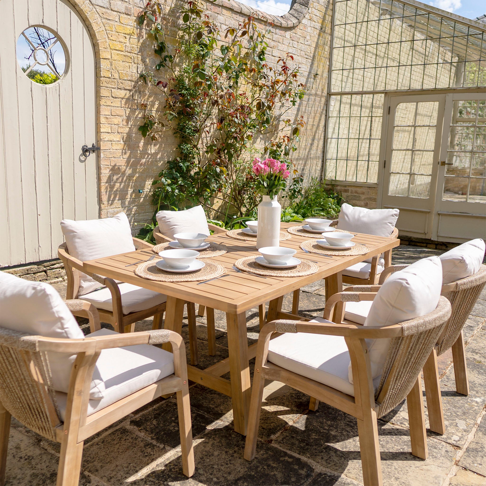Outdoor dining area featuring the Quay 6 Seat Rectangular Dining Set in Linen, with a sustainably sourced acacia wood table and cushioned chairs covered in water-resistant fabric, complemented by potted plants and a glass door in the background.
