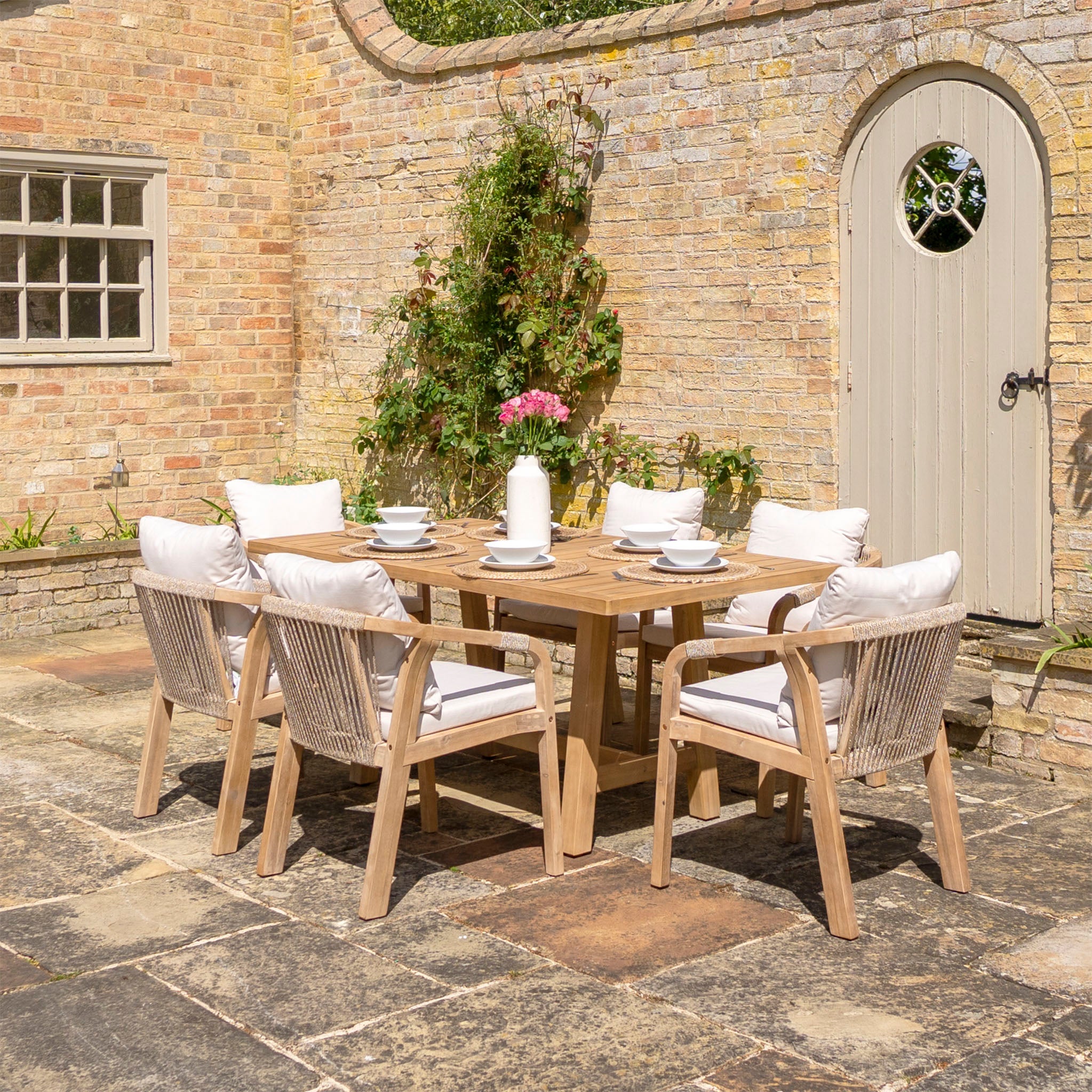 Outdoor patio featuring the Quay 6 Seat Rectangular Dining Set in Linen, comprised of a sustainably sourced acacia wood table and six cushioned chairs made with water-resistant fabric, complemented by elegant tableware and set against a brick wall backdrop with a white door and window.