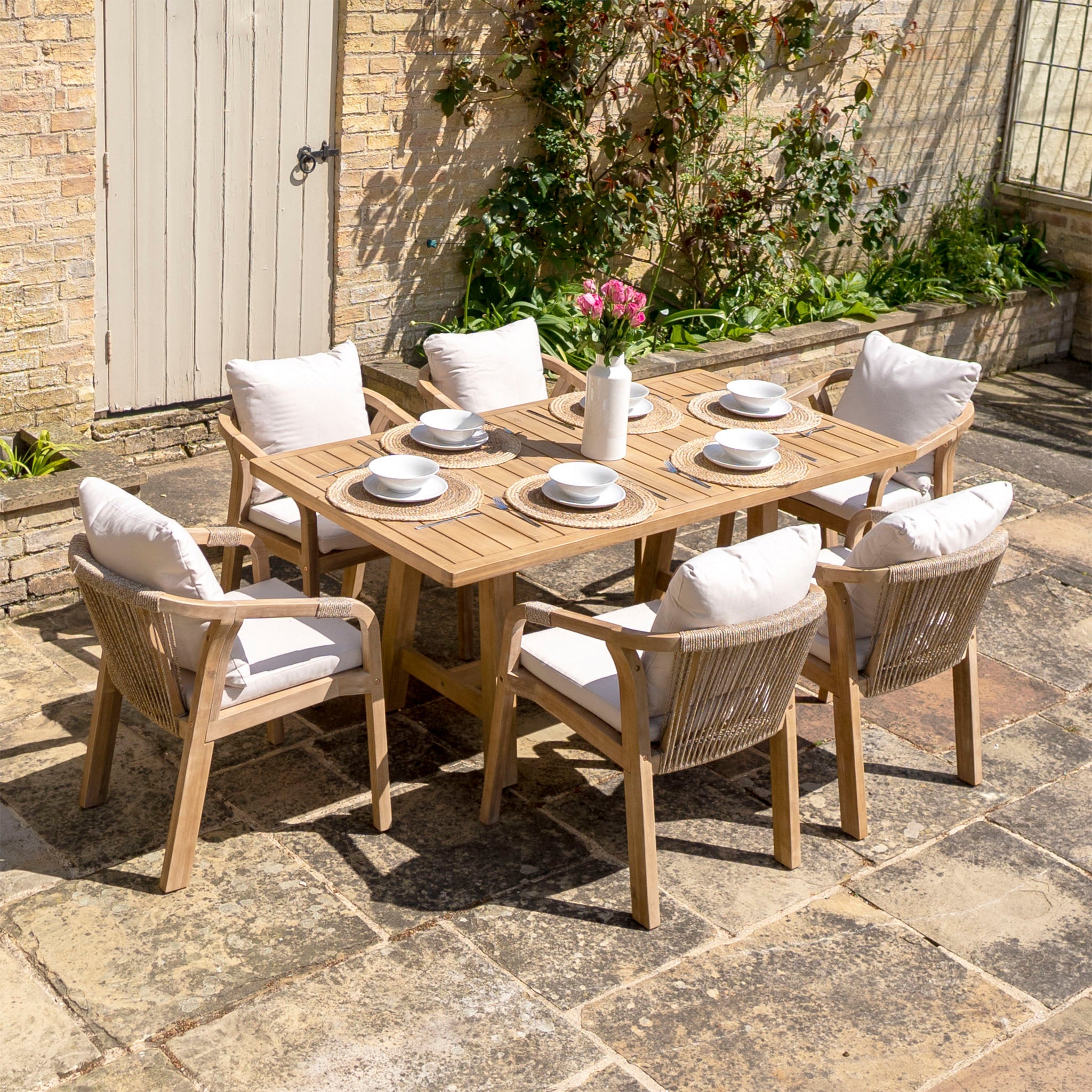 The Quay 6 Seat Rectangular Dining Set in Linen features six cushioned chairs wrapped in water-resistant fabric, surrounding a wooden table crafted from sustainably sourced acacia wood, and is elegantly set with white plates, cups, and a vase filled with pink flowers.