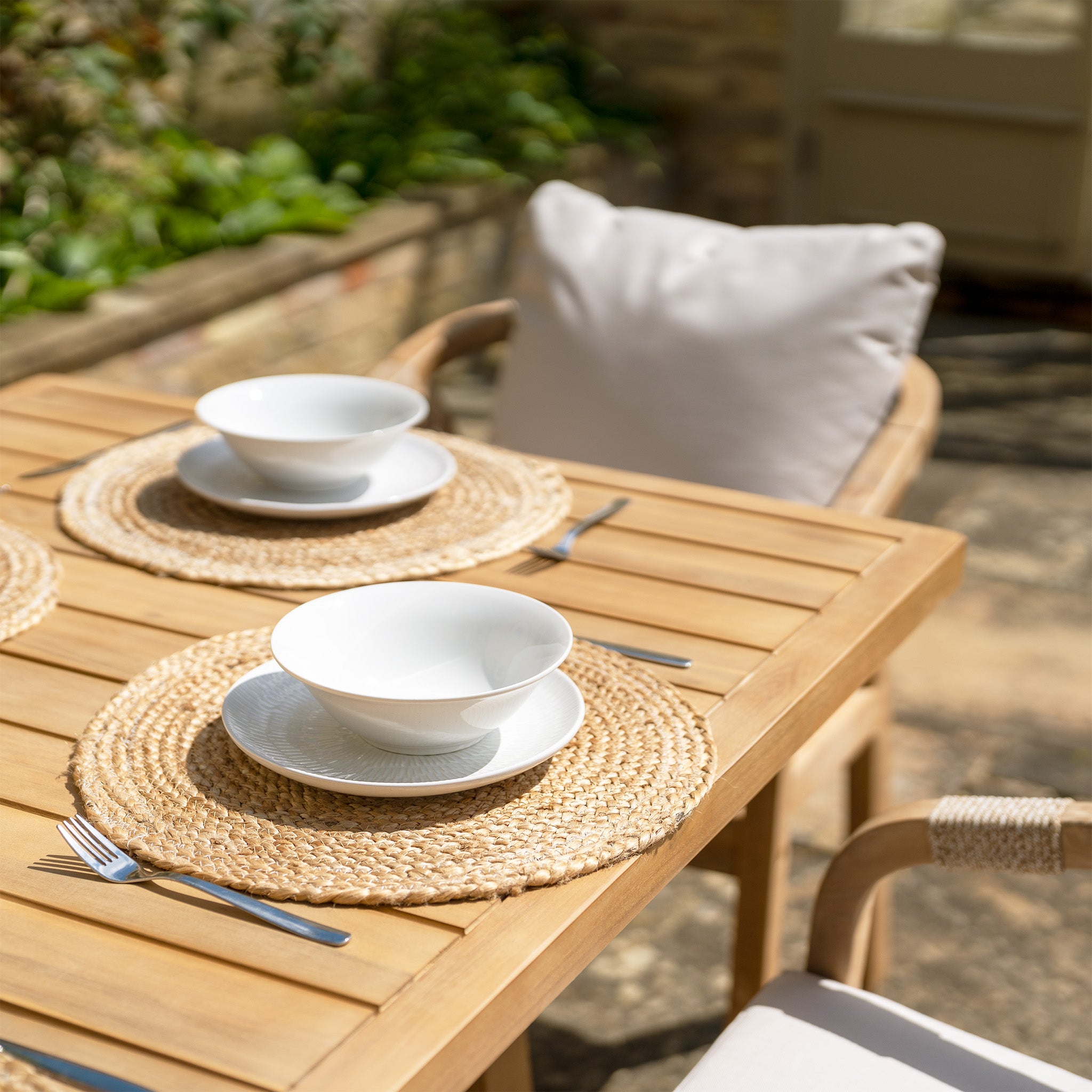 The Quay 6 Seat Rectangular Dining Set in Linen features a wooden outdoor dining setup with two place settings, including white bowls on plates, woven placemats, forks, and chairs adorned with water-resistant white cushions.