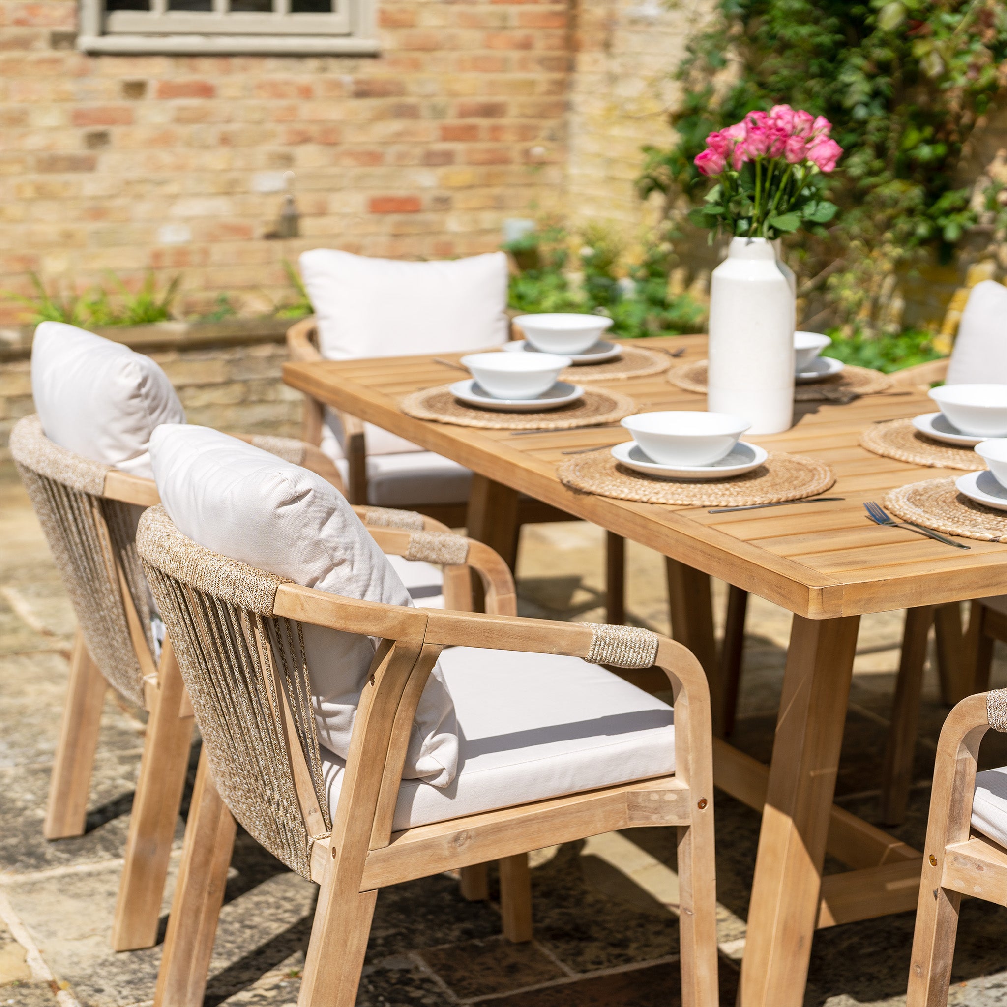 The Quay 6 Seat Rectangular Dining Set in Linen features a sustainably sourced acacia wood table, white cushioned chairs with water-resistant fabric, wicker placemats, white dishware, and a vase of pink flowers.