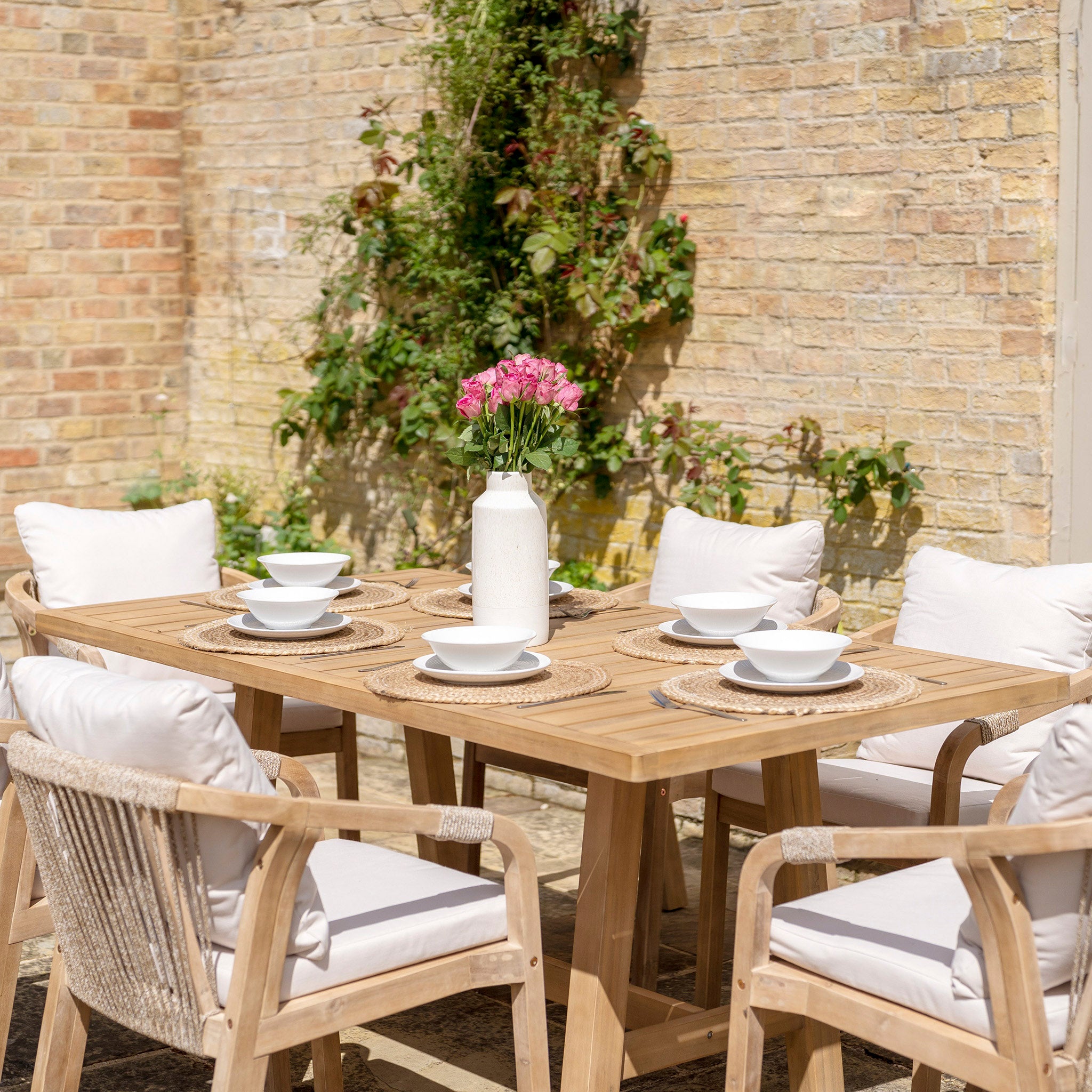 The Quay 6 Seat Rectangular Dining Set in Linen is made from sustainably sourced acacia wood and features six cushioned chairs with water-resistant fabric. This exquisite setup includes white dishes and a vase of pink flowers, all beautifully arranged against a brick wall.
