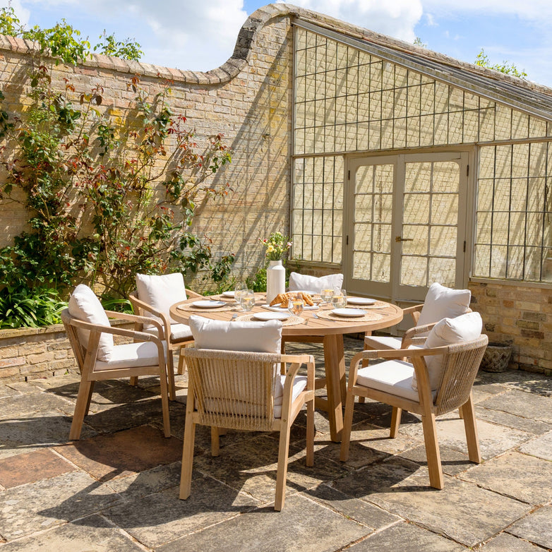 Transform your outdoor patio into an inviting retreat with the Quay 6 Seat Round Dining Set in Linen. This elegant setup includes a round wooden table crafted from sustainably sourced acacia wood, comfortable cushioned chairs upholstered in water-resistant polyester fabric, and a delightful dining area set against the backdrop of a brick wall adorned with climbing plants and a glass door.