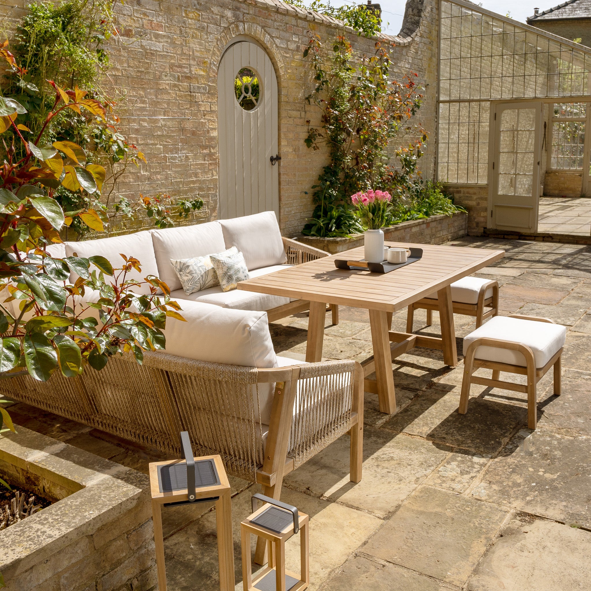 Outdoor patio featuring the Quay Corner Dining Set in Linen with a wooden table, cushioned bench, two chairs crafted from sustainably sourced acacia wood, and potted plants against a stone wall and garden door. The cushions are covered in water-resistant polyester fabric for added durability.