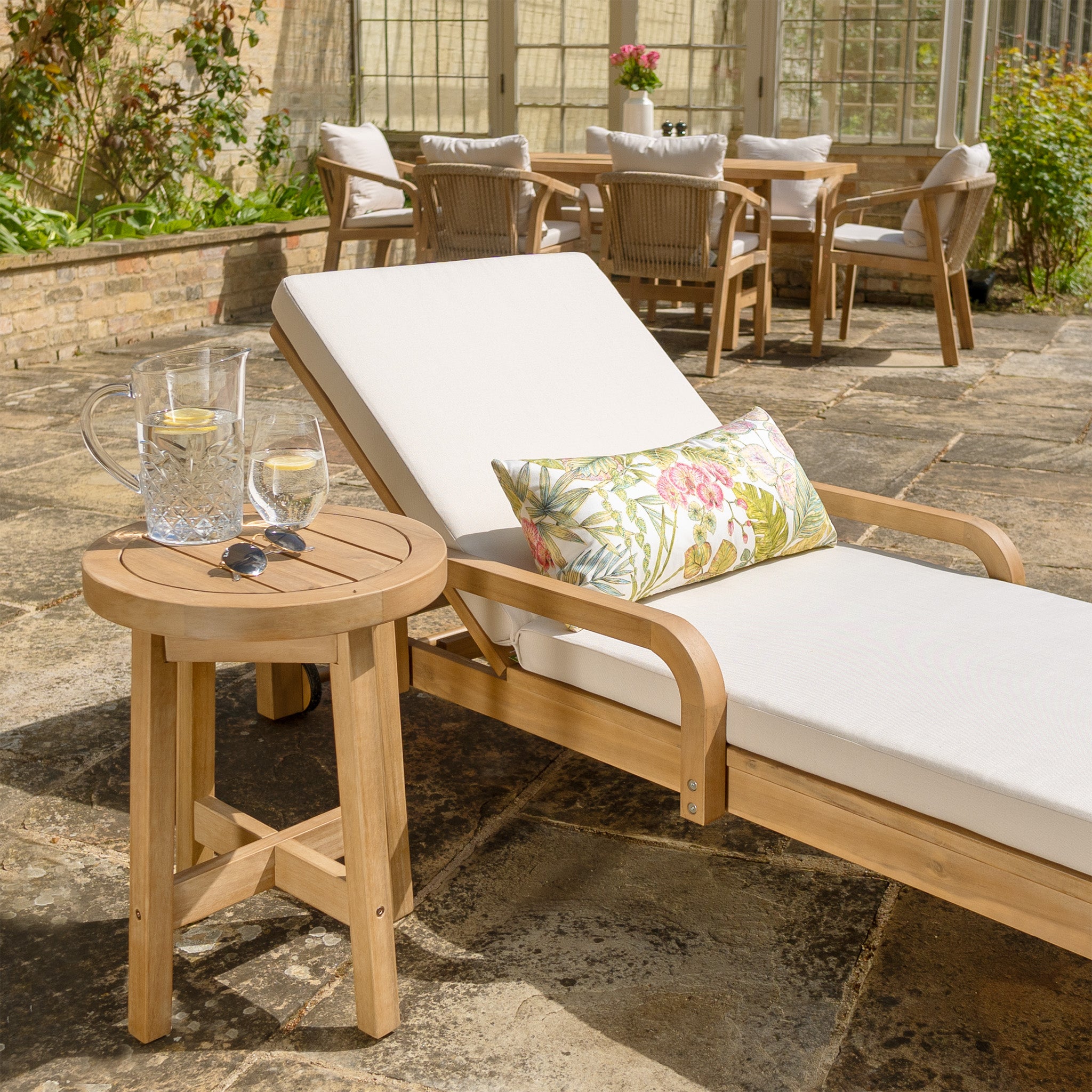 Quay Sun Lounger In Linen from the Quay collection with a tropical pillow beside a small table holding drinks and sunglasses, surrounded by outdoor furniture made of acacia wood.