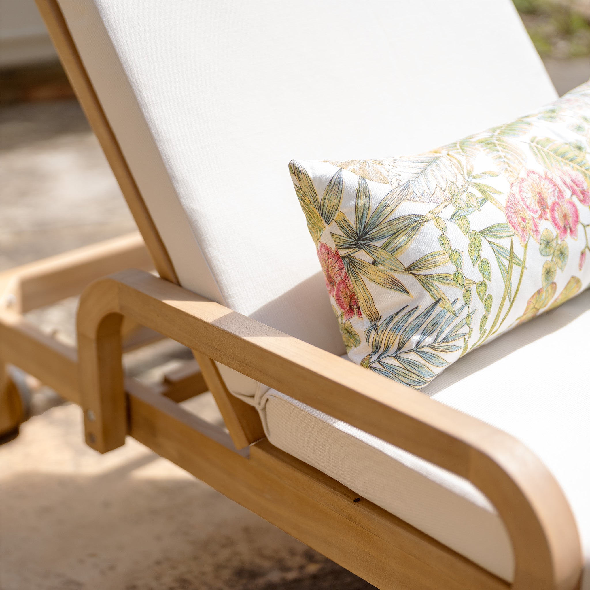 A Quay Sun Lounger in Linen, crafted from acacia wood with white cushions and adorned with a floral-patterned pillow, set outdoors on a sunny day.