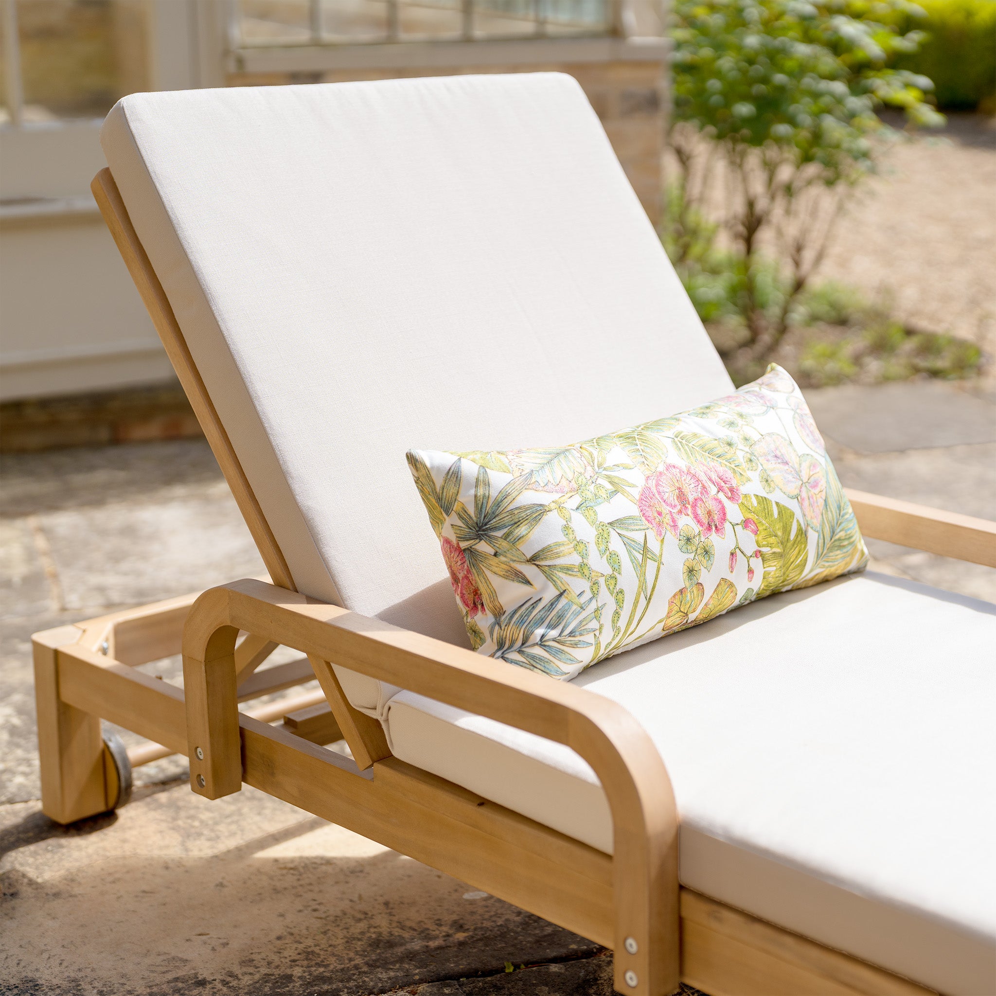 A Quay Sun Lounger in Linen, adorned with white cushions and a floral pillow, sits on a stone patio near lush greenery.