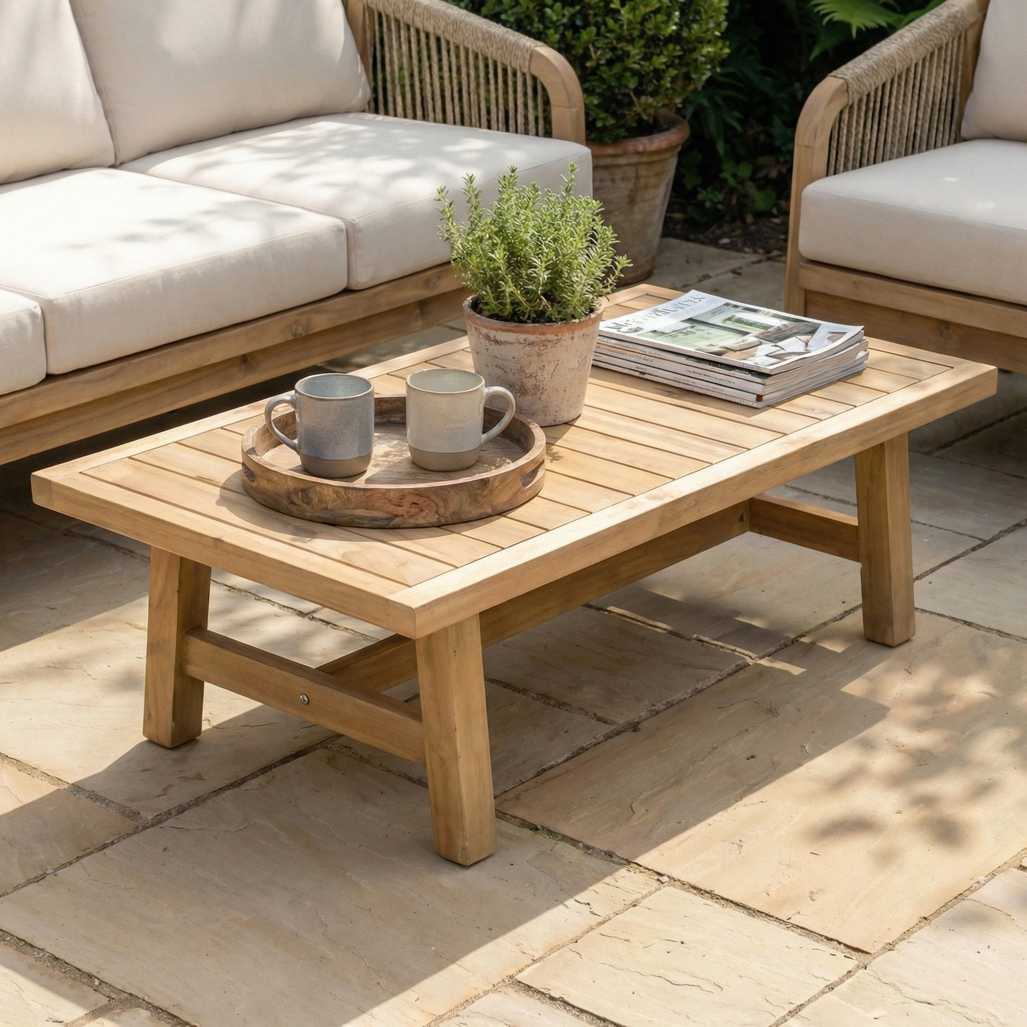 The Quay 3 Seat Sofa Set in Linen features a wooden patio table with mugs, a plant pot, and magazines, arranged between outdoor lounge chairs for a stylish garden setup on a stone patio.