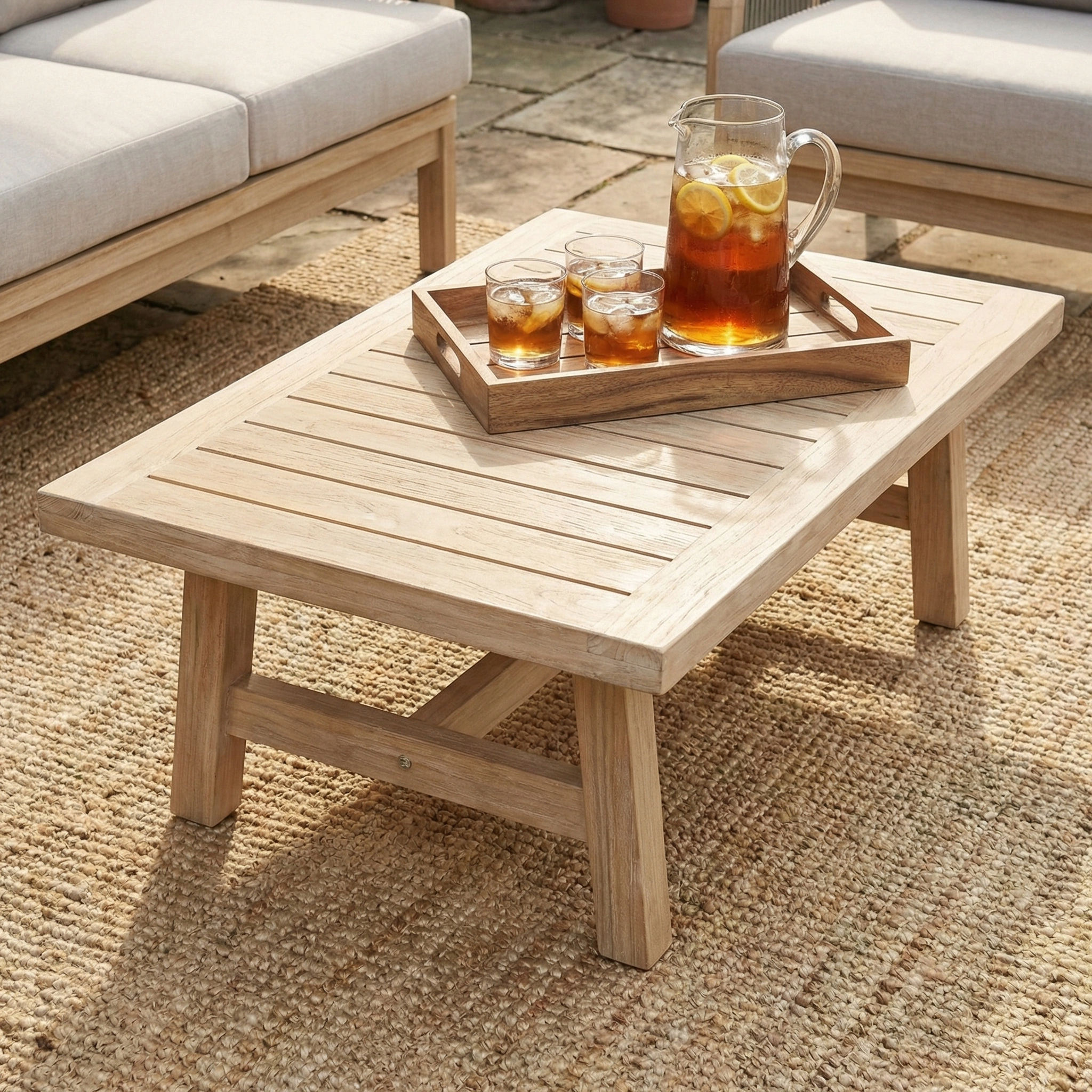 A Quay 3 Seat Sofa Set in Mocha rests on a woven outdoor rug, with a wooden patio table nearby holding a tray with a pitcher and two glasses of iced tea.