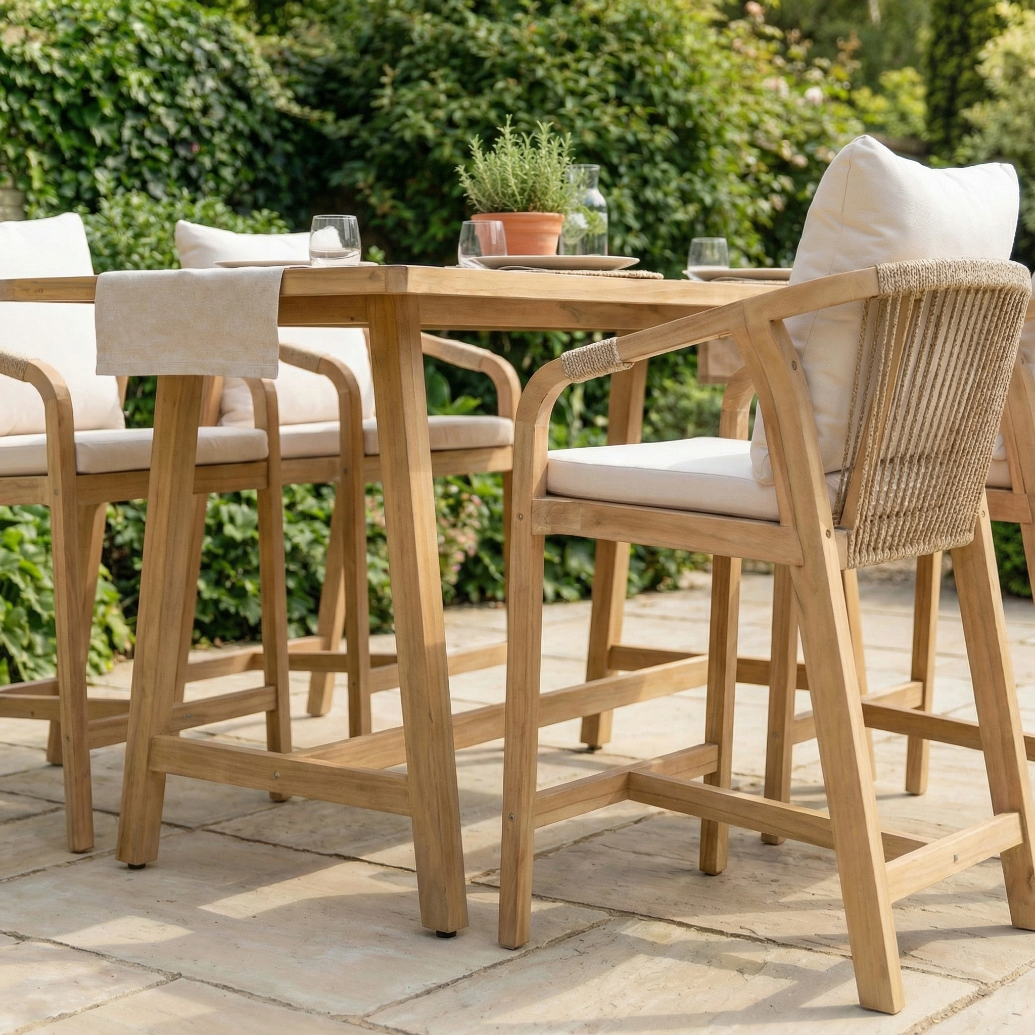 The Quay 4 Seat Rectangular Bar Set in Linen features an acacia wood table and chairs with white cushions, arranged on a stone terrace and surrounded by lush greenery.