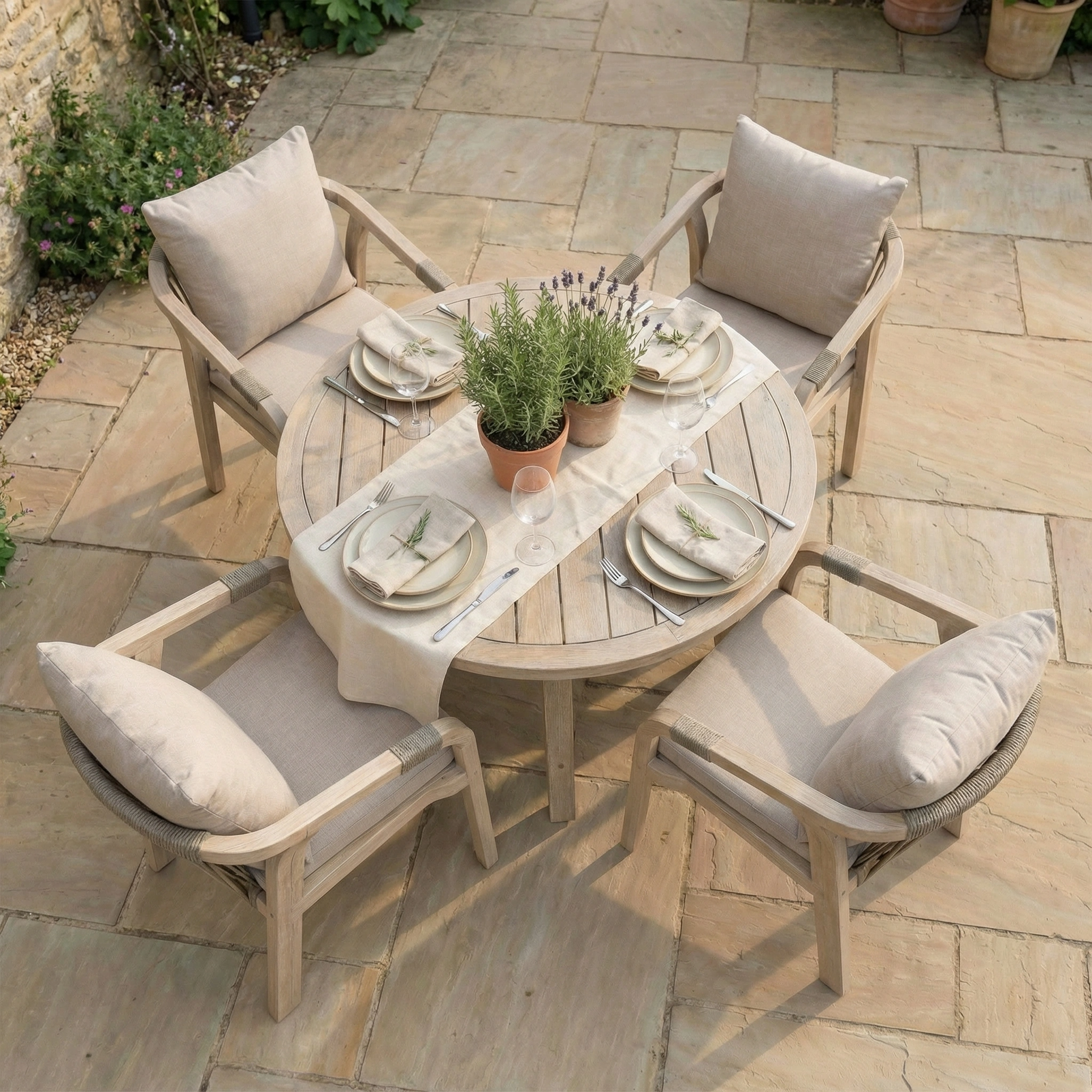 The Quay 4 Seat Round Dining Set in Mocha features a round eucalyptus wood table and four cushioned chairs, set with plates, cutlery, and potted herbs—ideal patio furniture for outdoor dining and gatherings.