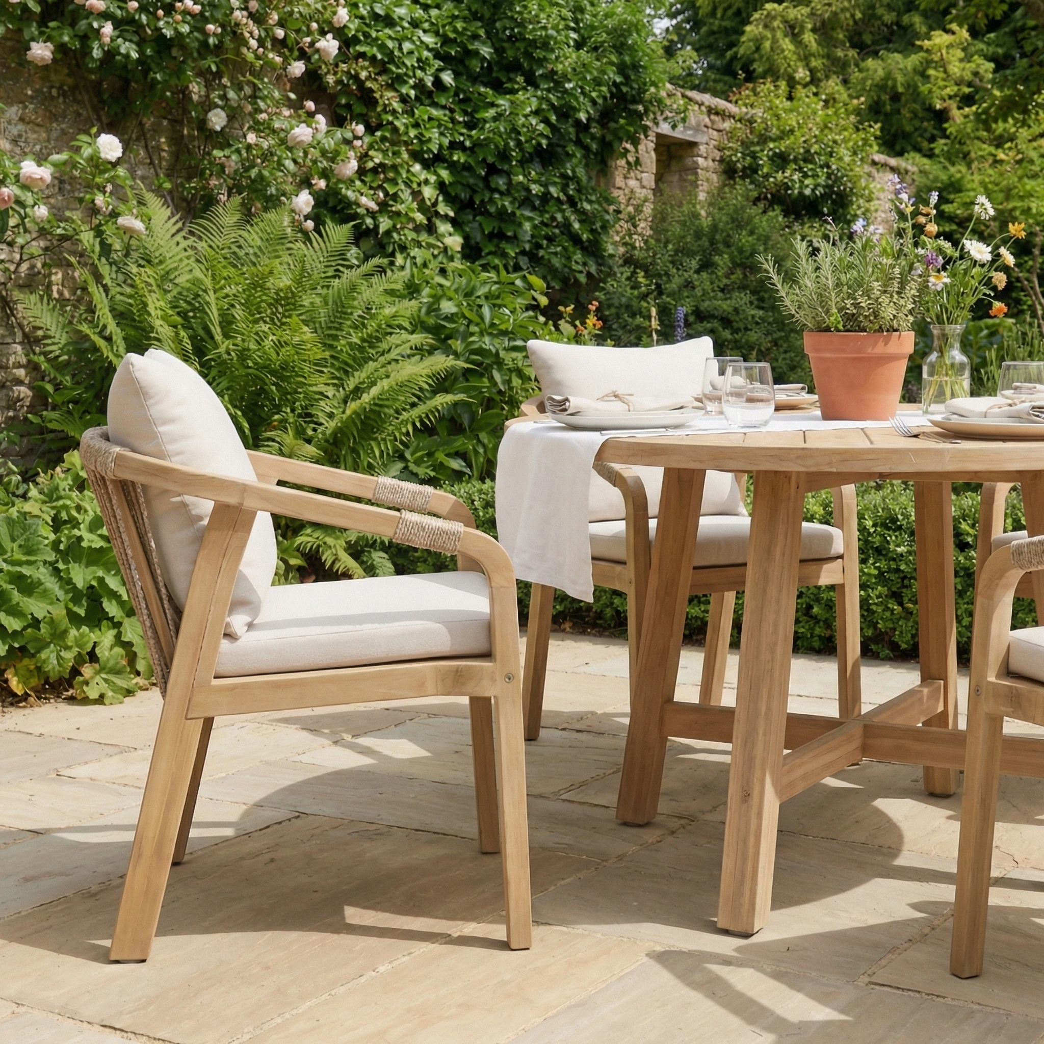 The Quay 4 Seat Round Dining Set in Linen features rope backrest chairs with cushions, ideal for outdoor dining amid garden greenery and flowers.