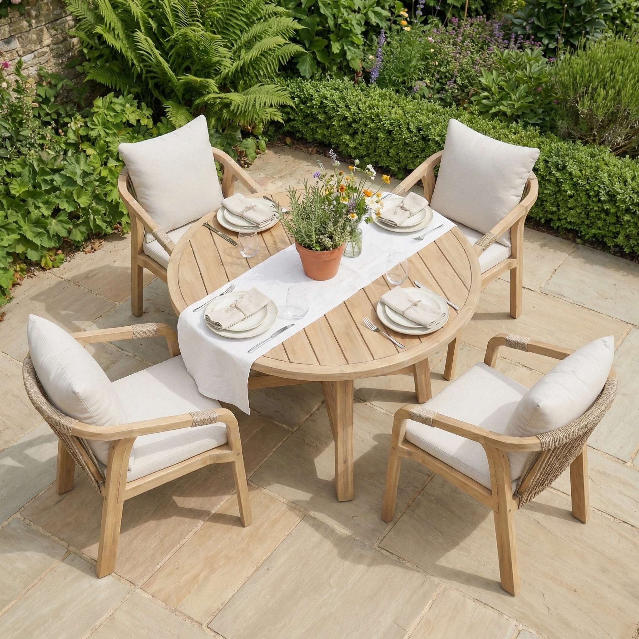 The Quay 4 Seat Round Dining Set in Linen features rope backrest chairs and a round wooden table, styled outdoors with plates and a potted plant centerpiece.