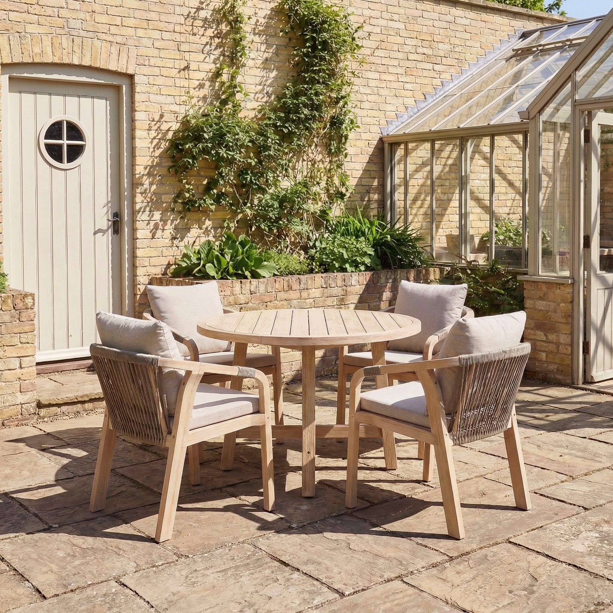 The Quay 4 Seat Round Dining Set in Mocha includes a wooden patio table and four cushioned chairs, ideal for outdoor spaces like patios near greenhouses or ivy-covered brick walls.