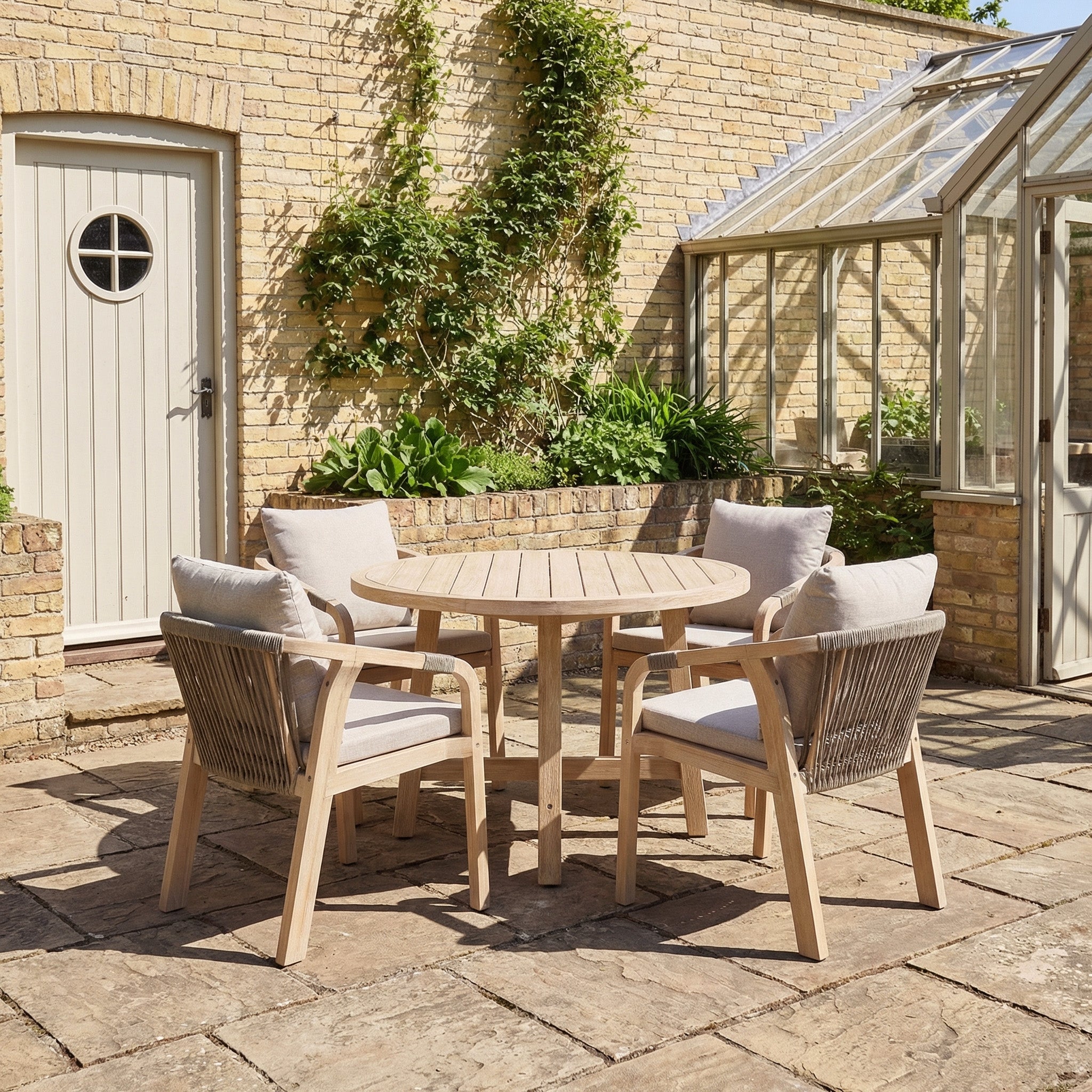 The Quay 4 Seat Round Dining Set in Mocha, with cushioned chairs, sits on a stone patio by a brick wall and glass greenhouse, offering an inviting space for outdoor dining and gatherings.
